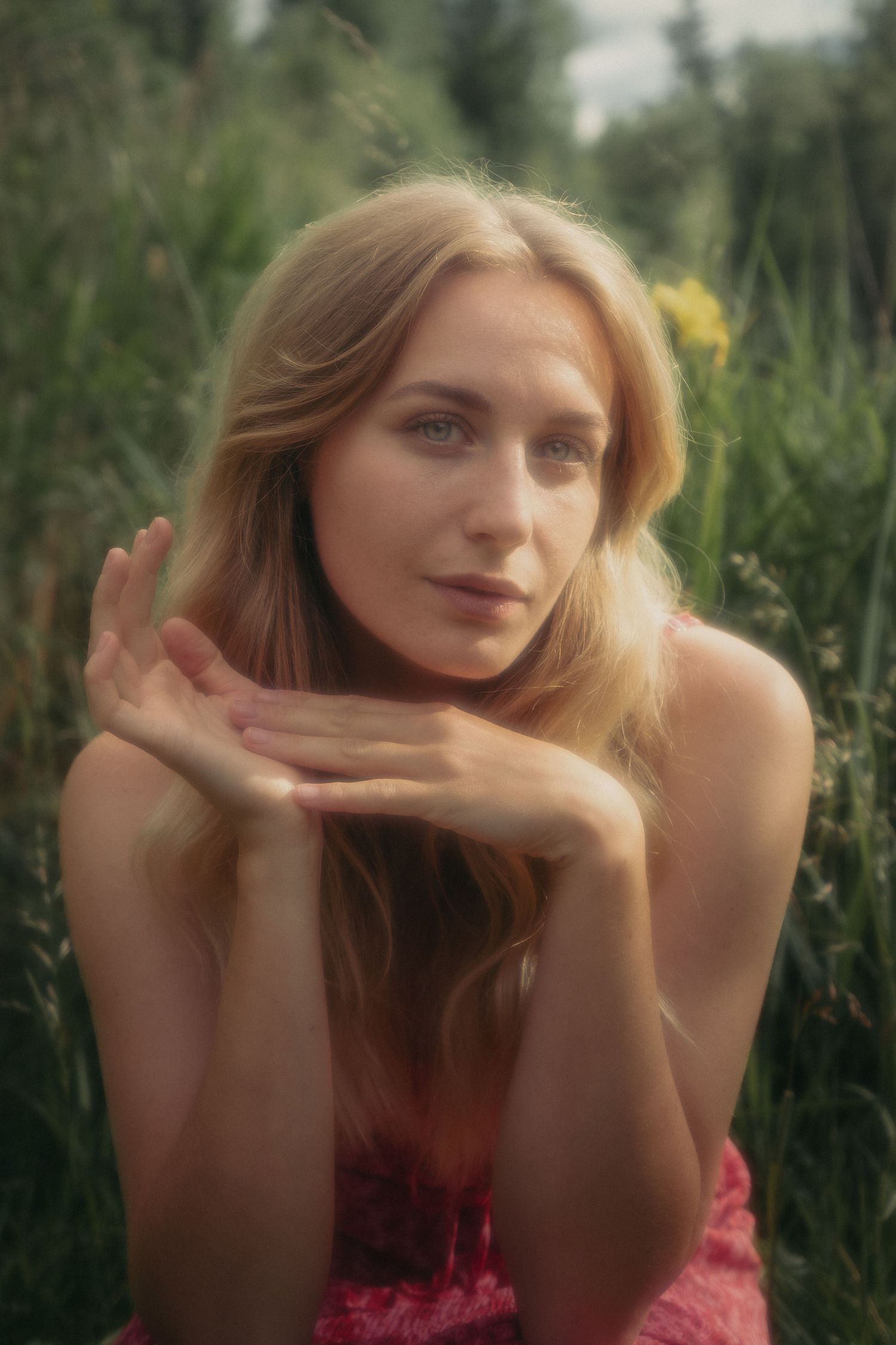Blonde woman in pink dress with blue eyes posing gracefully in green meadow.