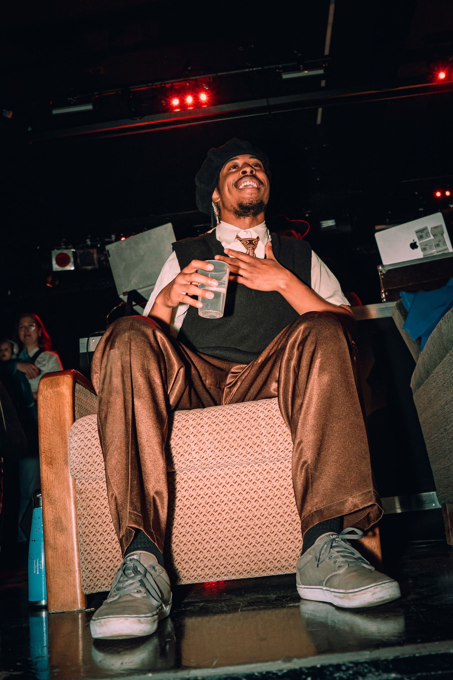 A smiling man in a beanie and brown pants sits in a chair holding a drink under colorful stage lights.