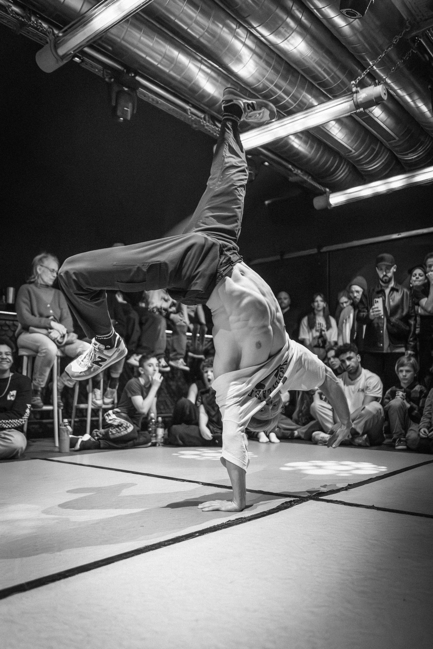 Shirtless breakdancer performs a one-handed handstand freeze at an indoor b-boy competition.