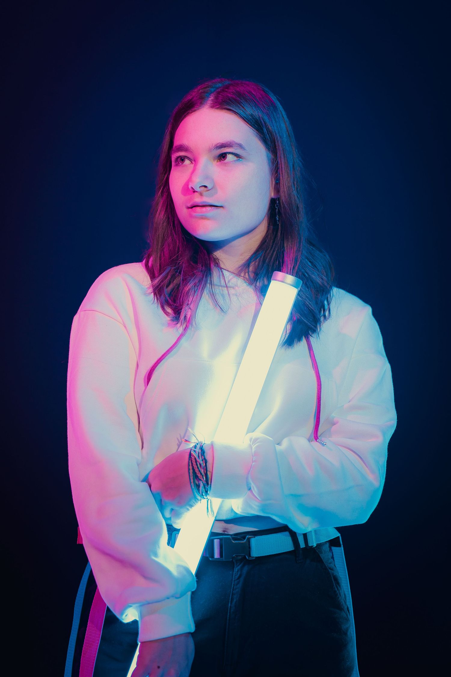 Young woman holding a glowing neon tube in a dark studio with pink and blue lighting.