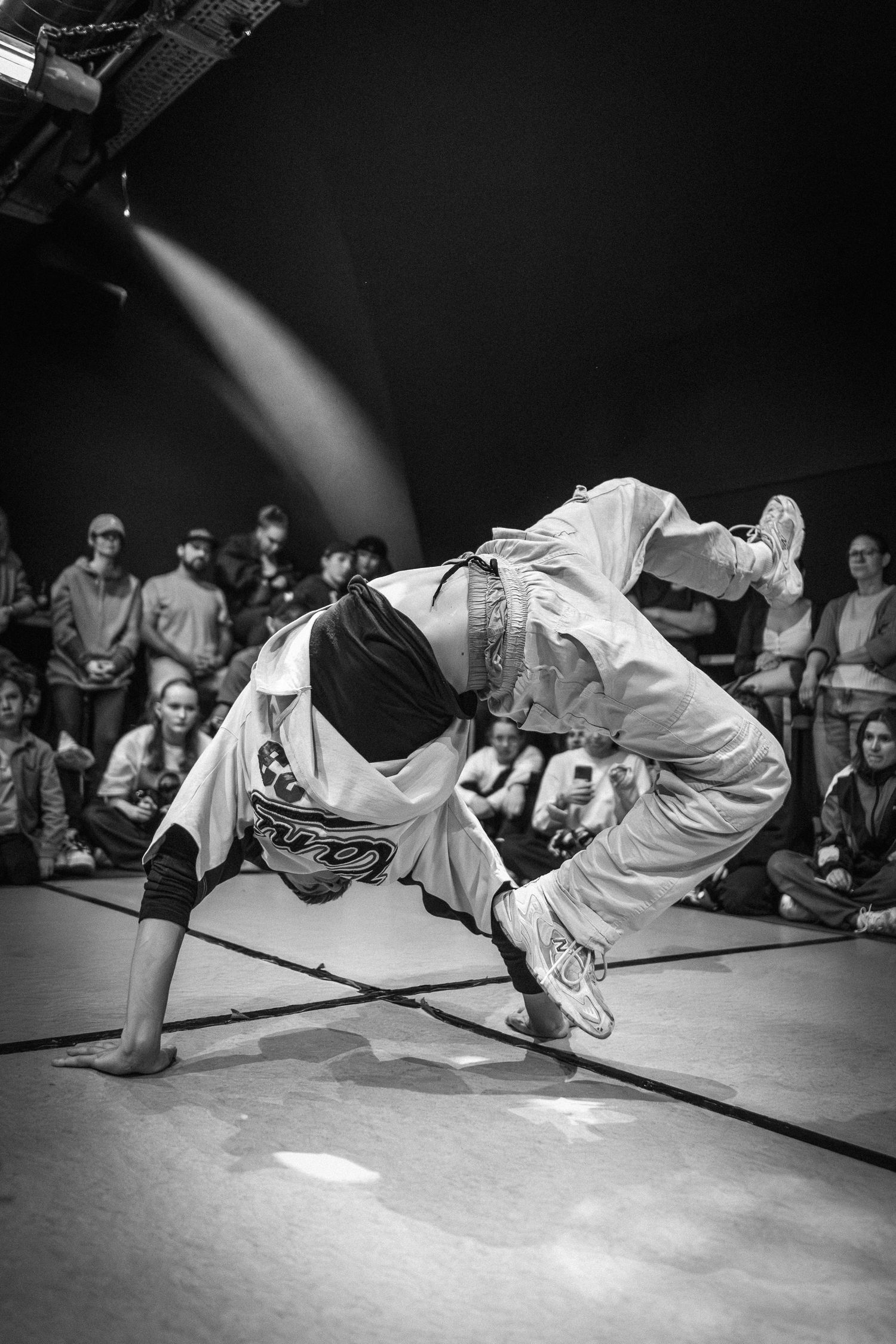 B-boy executing a dynamic freeze move at an indoor breakdancing competition in black and white.