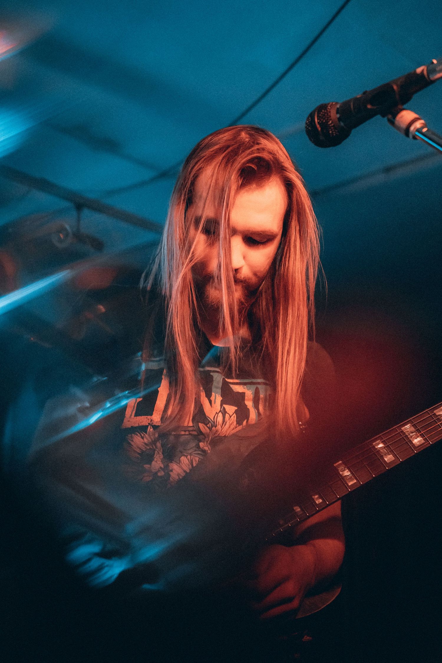 Long-haired guitarist playing electric guitar under warm red and blue stage lights.