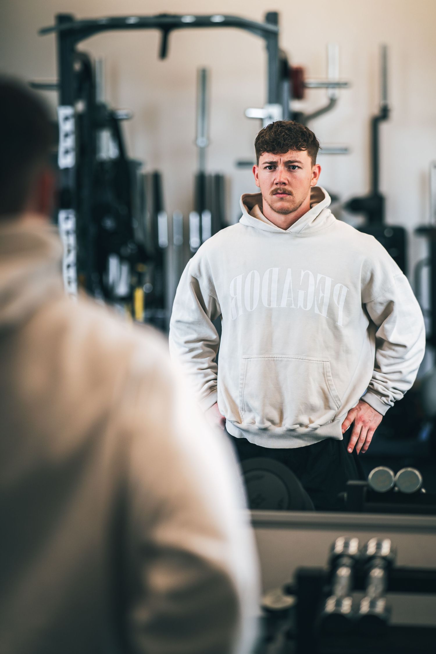 Young man in gray Reebok hoodie standing in gym looking in mirror with workout equipment in background.