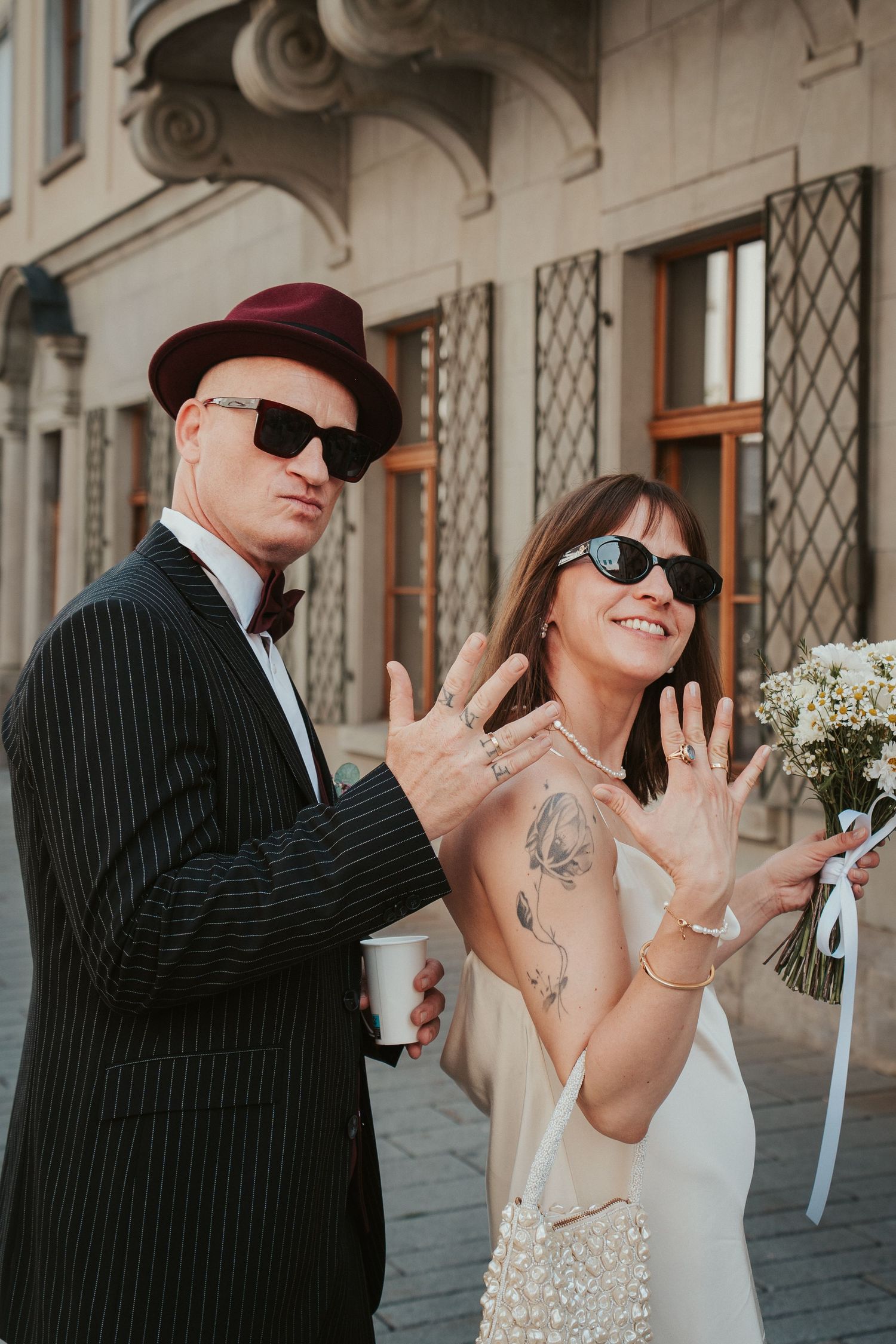 Stylish newlyweds in sunglasses showing off wedding rings, groom in pinstripe suit with hat, bride holding bouquet.