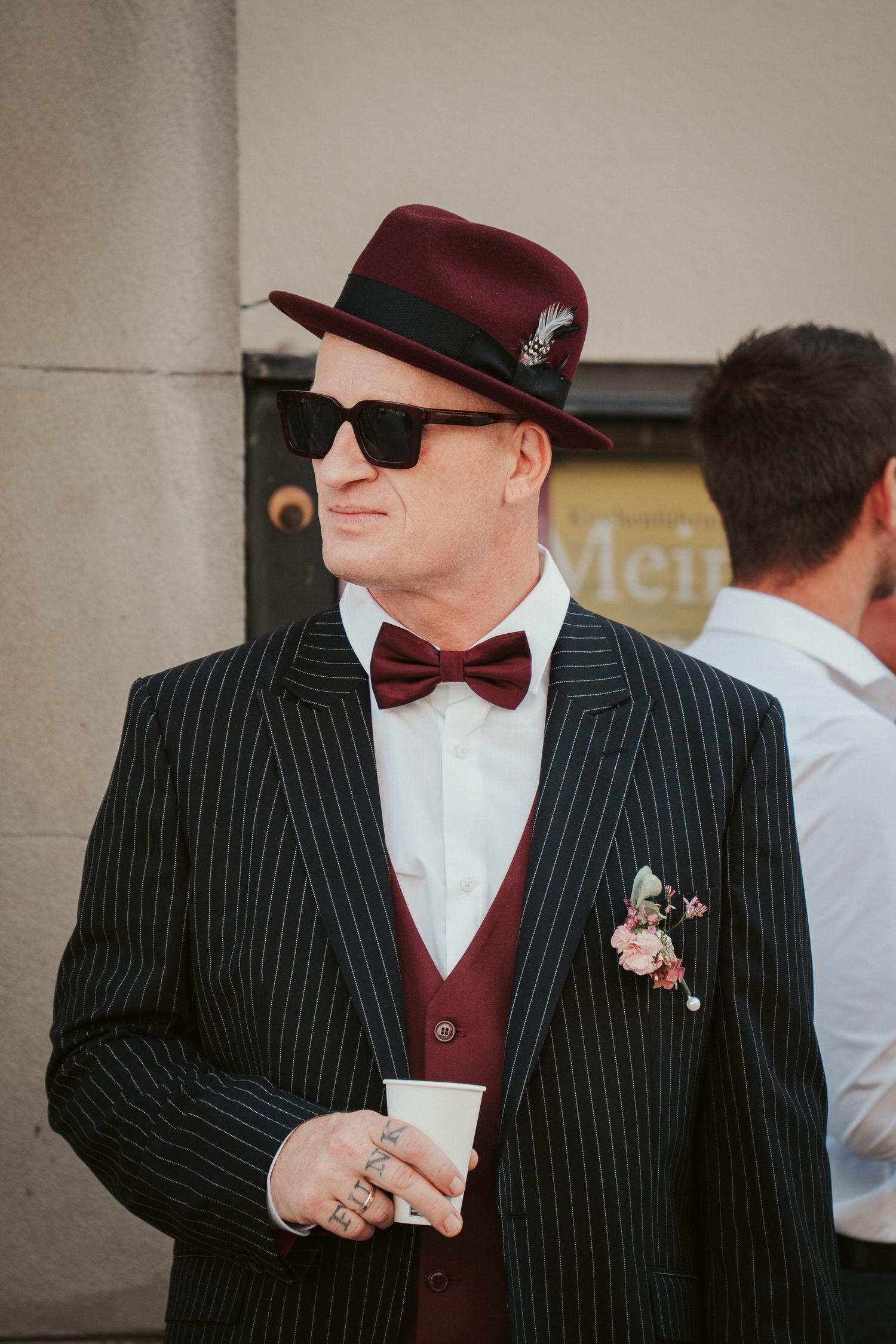 A stylish groom in a pinstripe suit, burgundy hat, bow tie, and sunglasses holds a cup at an outdoor wedding.