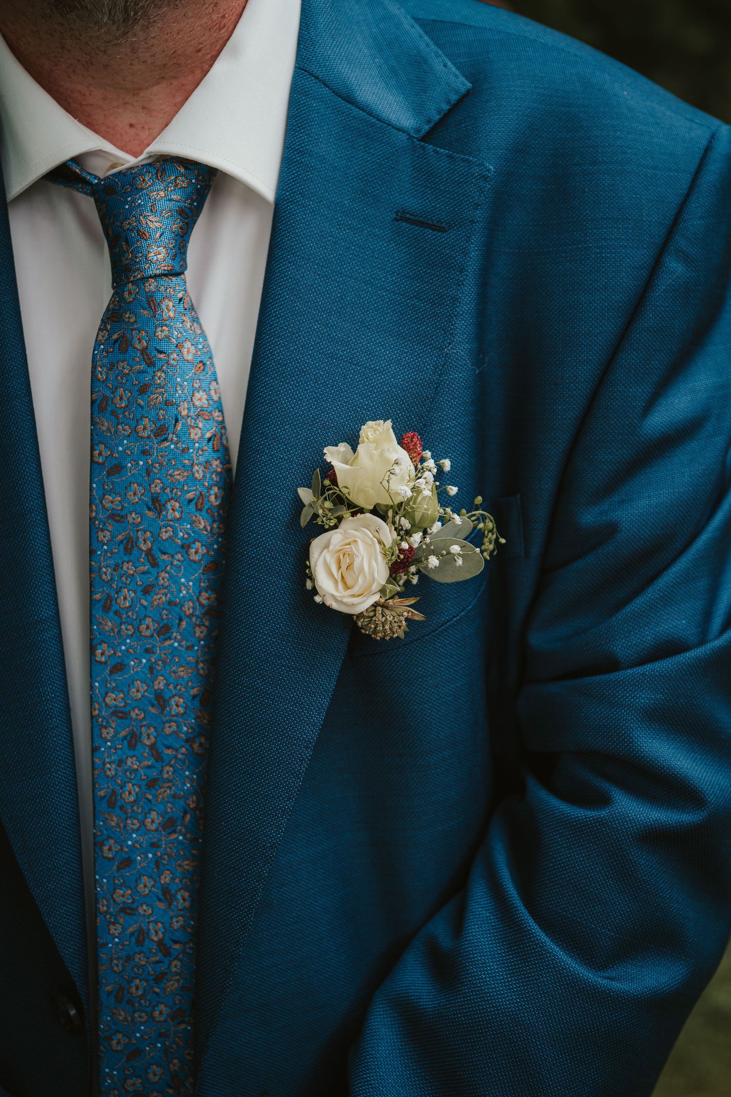 Close-up of groom in blue suit with patterned tie and white rose boutonniere on lapel.