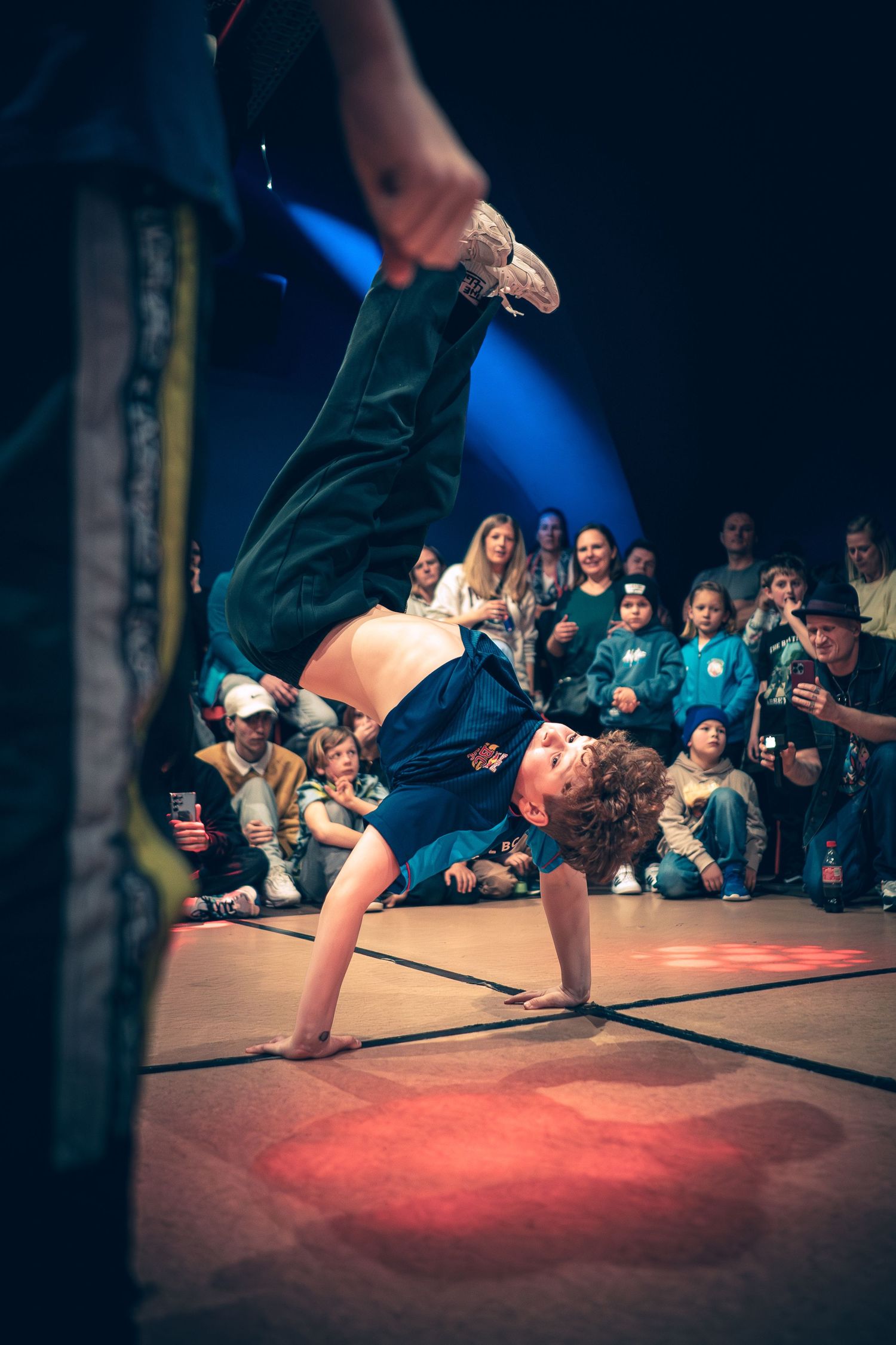 Young curly-haired breakdancer performs a headstand freeze at a vibrant b-boy battle competition.