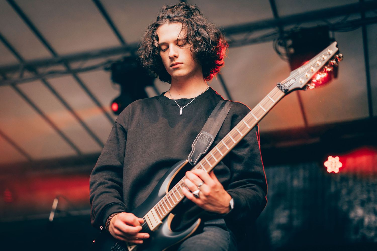 Young guitarist with curly hair plays electric guitar on stage under red concert lighting.