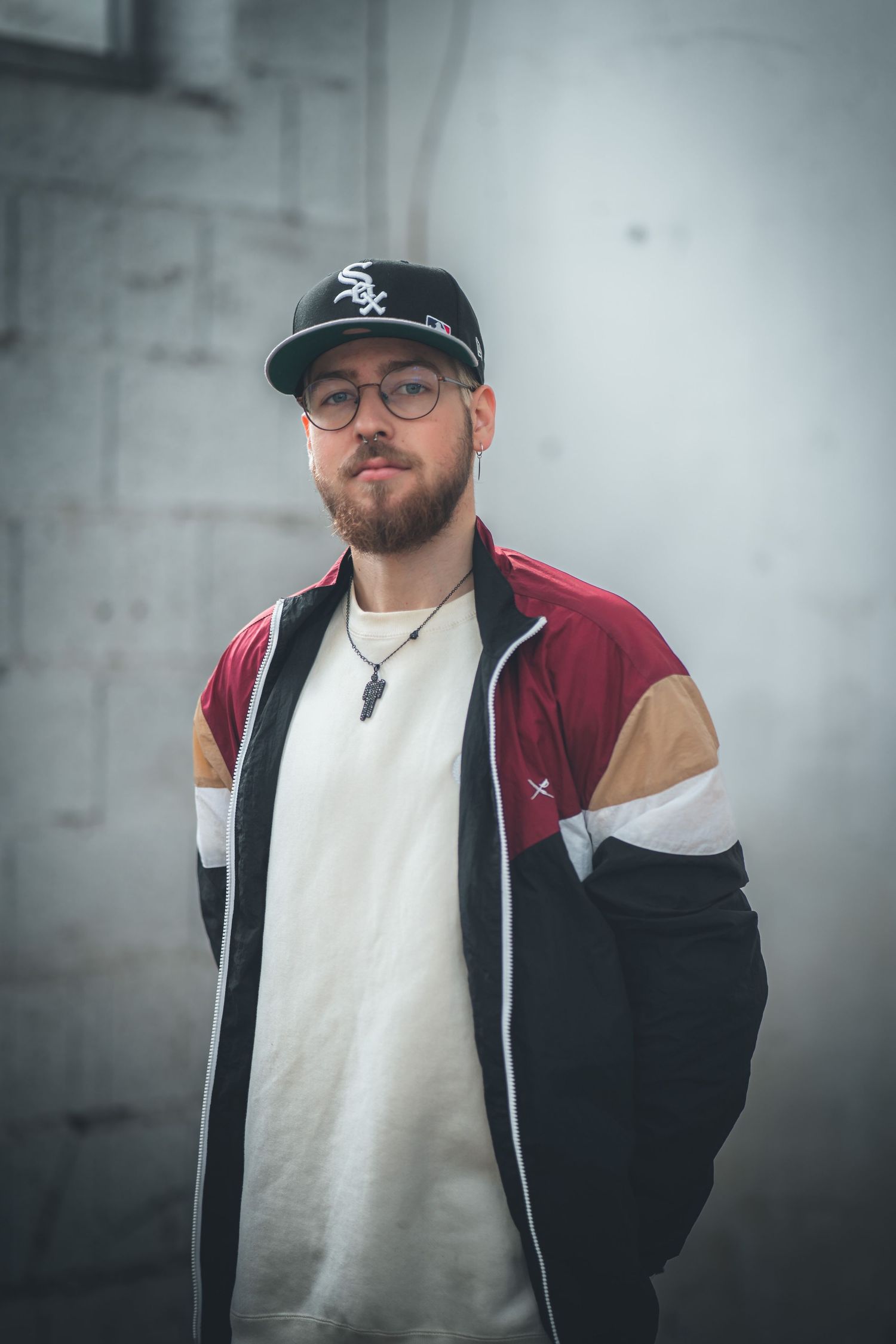 Bearded man wearing Chicago White Sox cap and colorblock jacket posing against concrete wall.