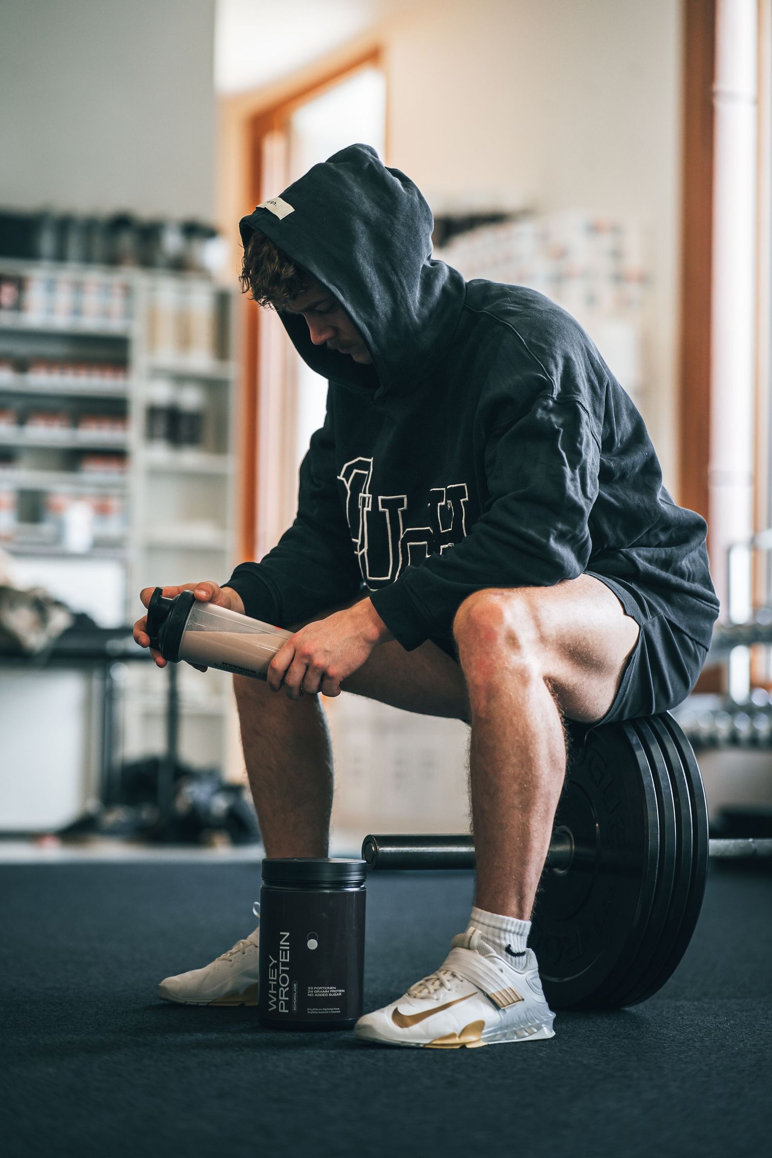 Athlete in black hoodie sitting on weight plates holding a shaker bottle next to supplement tub.
