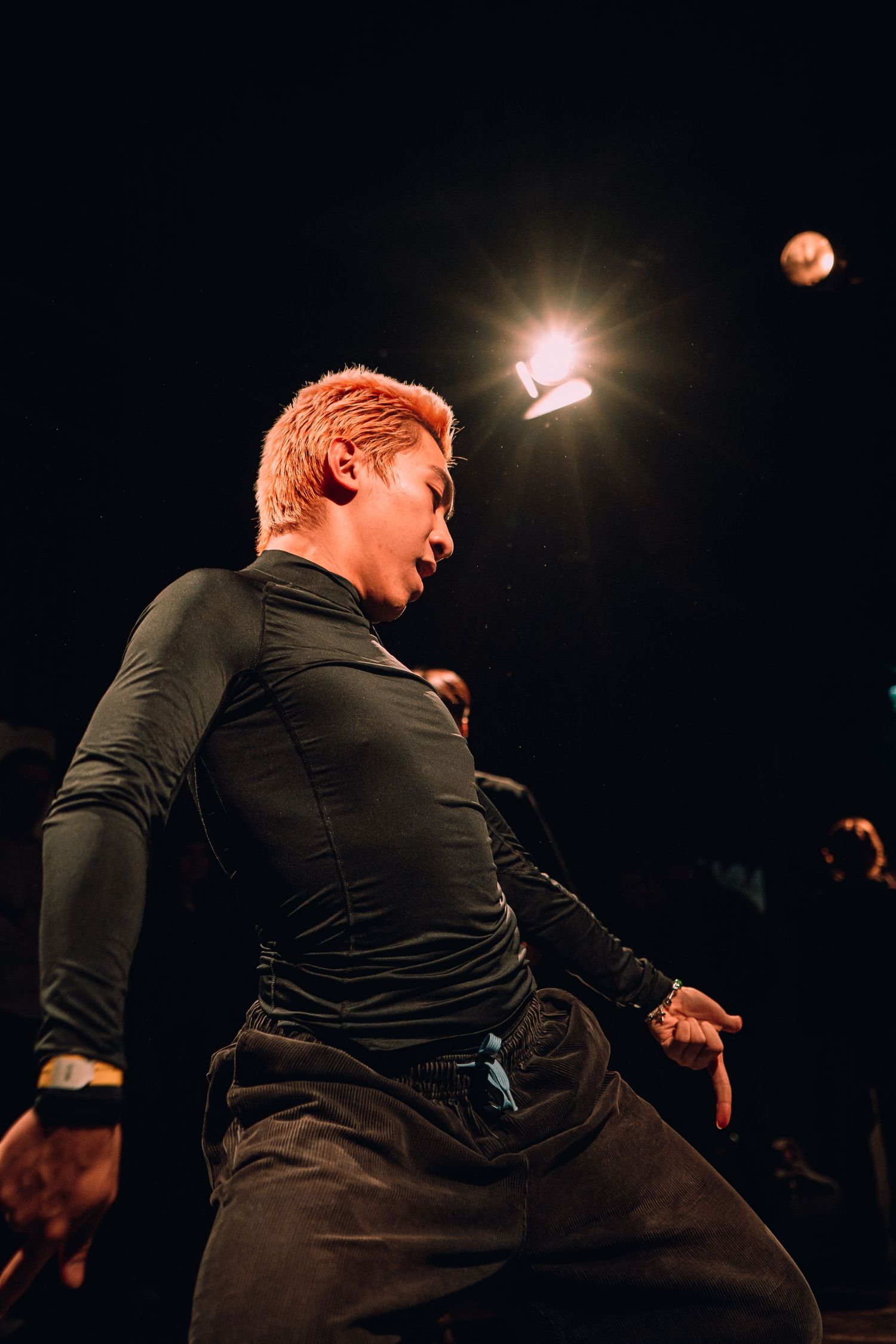 A performer with orange hair dances energetically on stage under a bright spotlight in a dark venue.