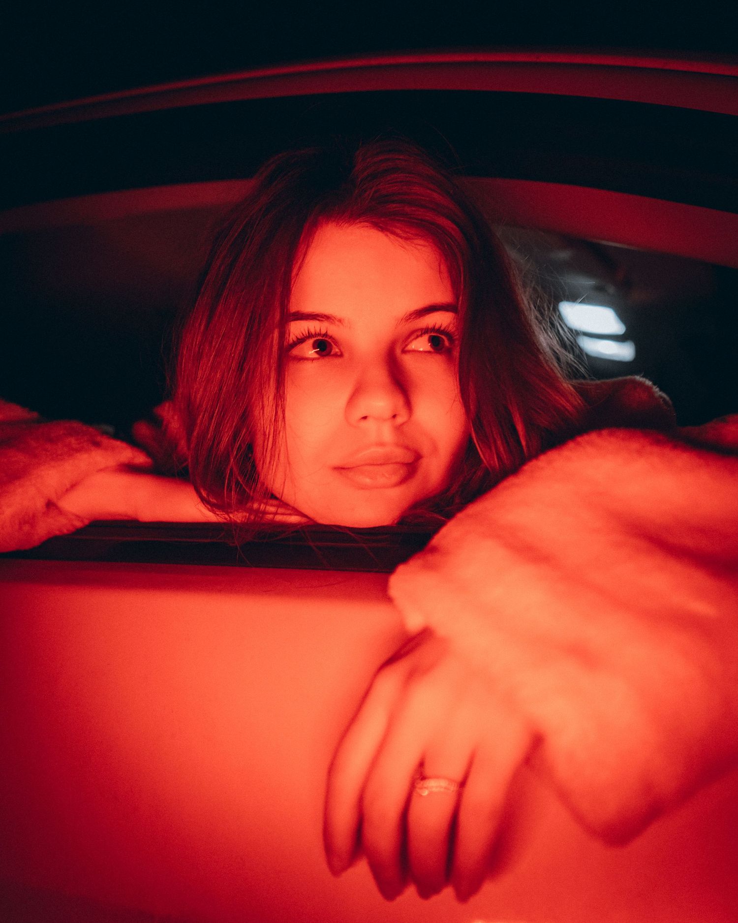 Portrait of a young woman resting her arms on a car window under vivid red ambient lighting.