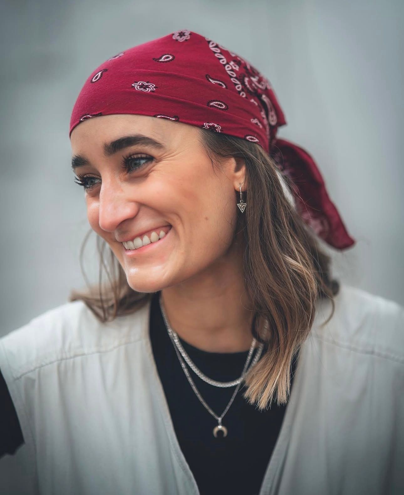 Smiling woman wearing a red paisley bandana headscarf with layered necklaces outdoors.
