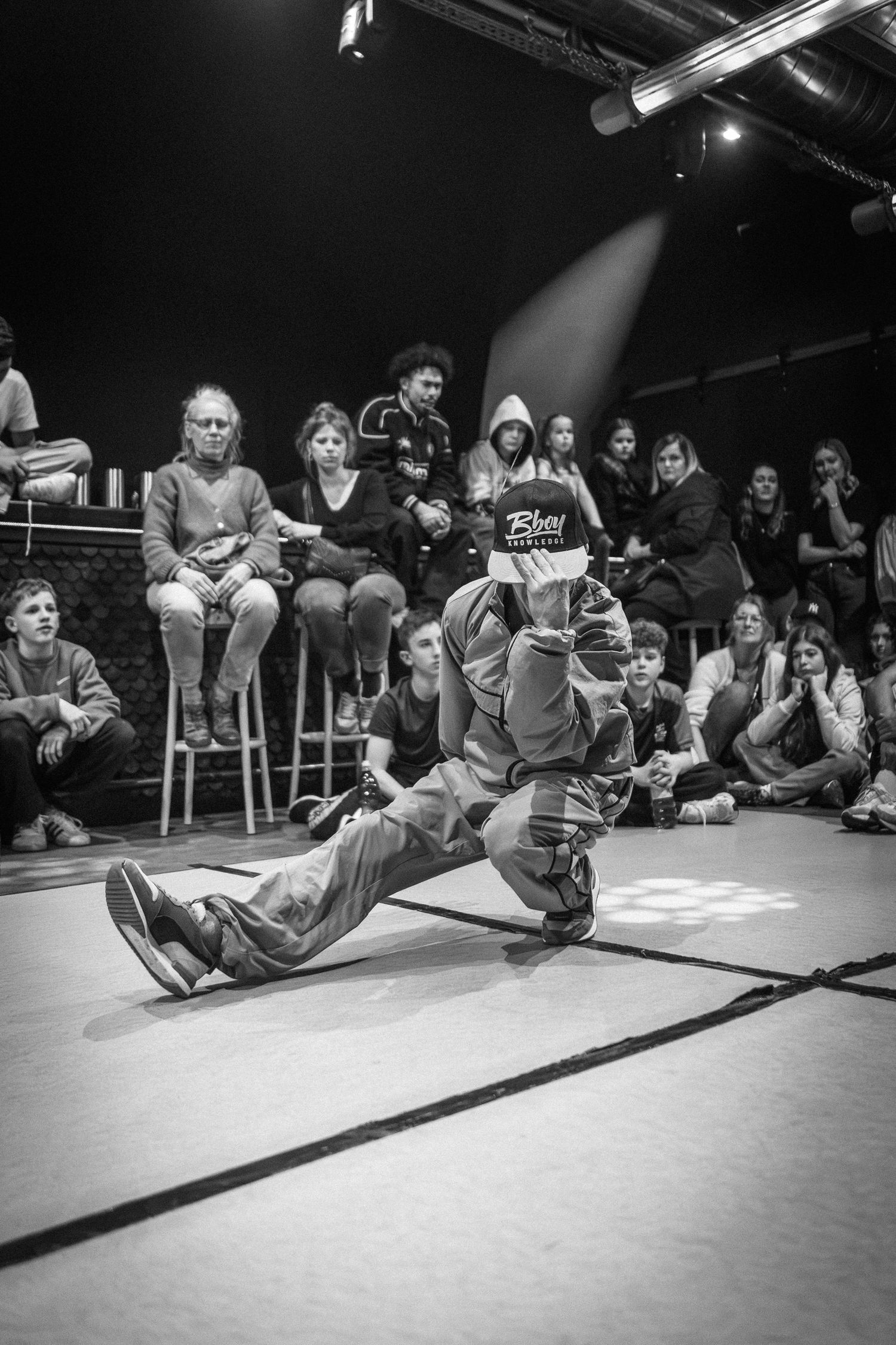 A breakdancer performs a low ground move on the floor surrounded by an engaged seated audience in black and white.