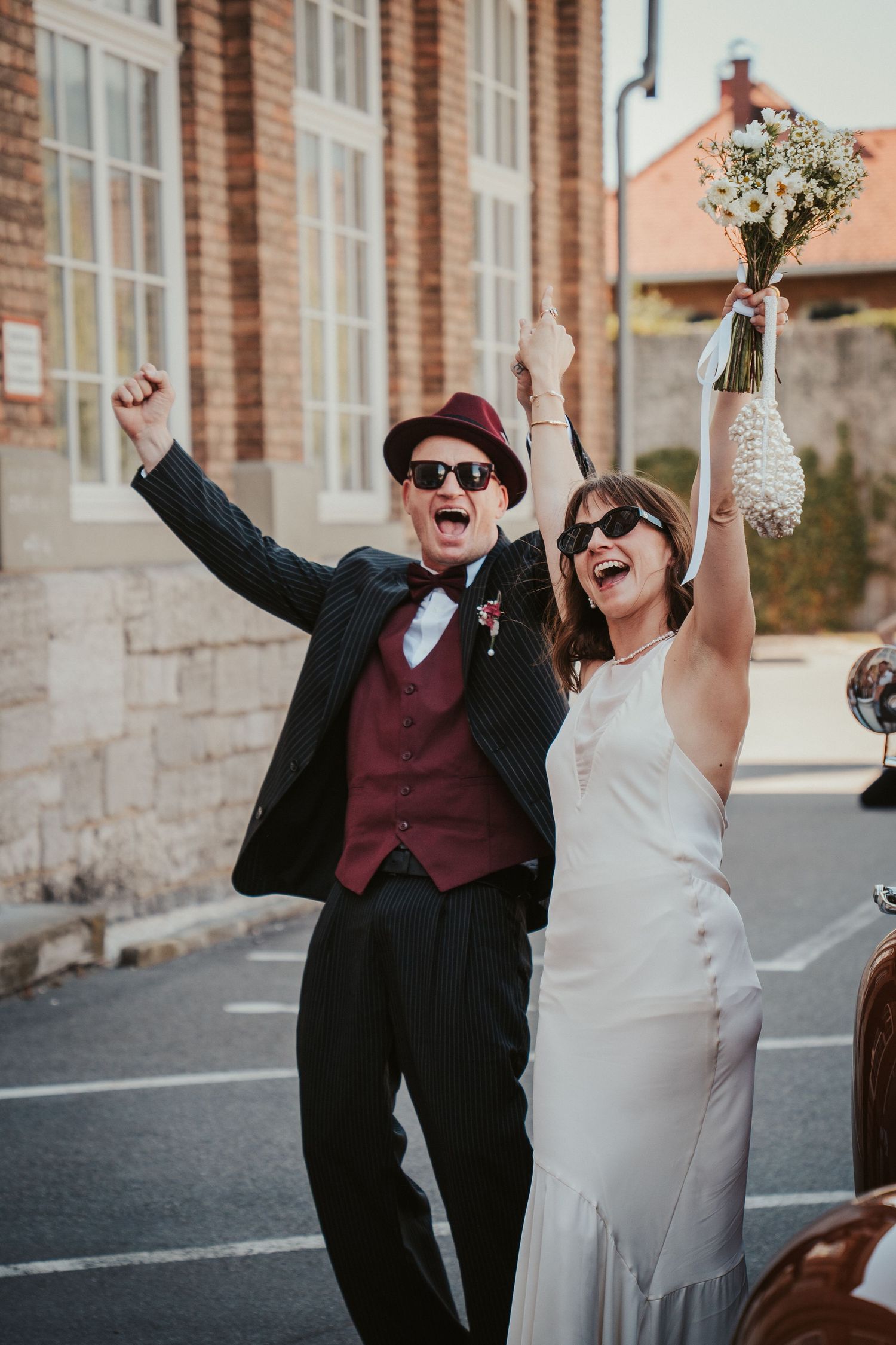 An ecstatic bride and groom in sunglasses celebrate joyfully outside with raised arms after their wedding ceremony.