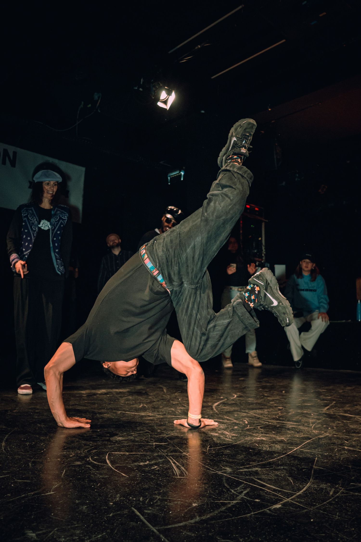 A breakdancer performs a one-handed freeze move on a dark stage while spectators watch in the background.