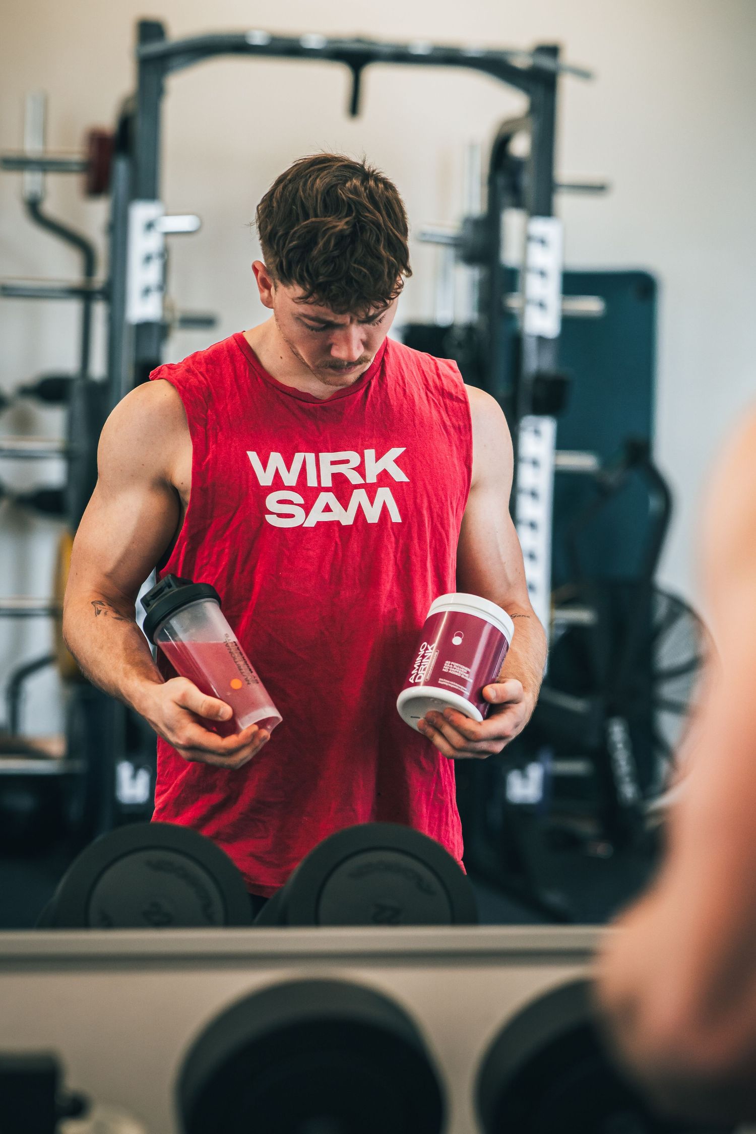 Muscular man in a red WIRK SAM tank top holds supplements and a shaker bottle in a gym.