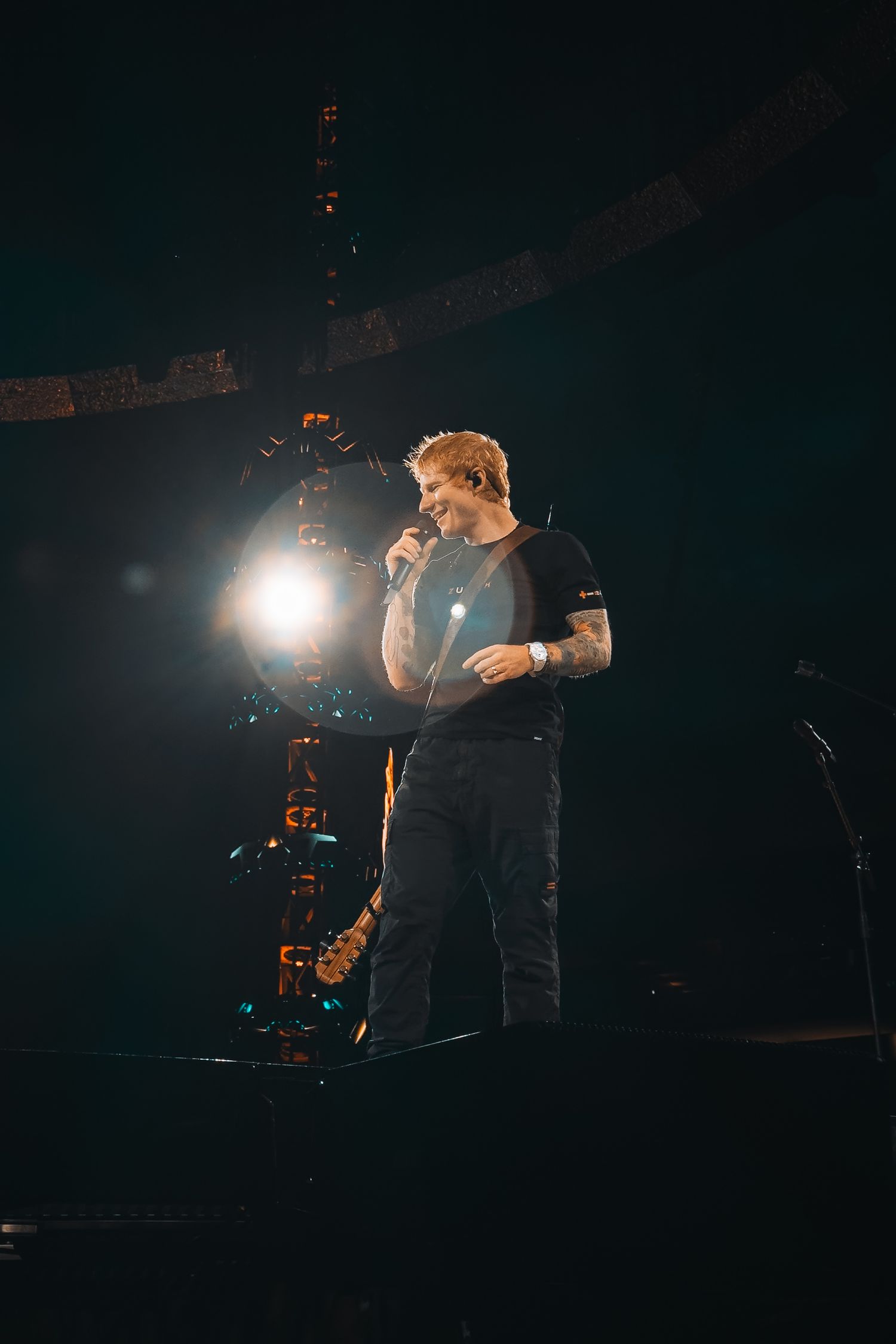 A red-haired male performer stands on a dark stage holding a microphone under bright spotlights at a live concert.