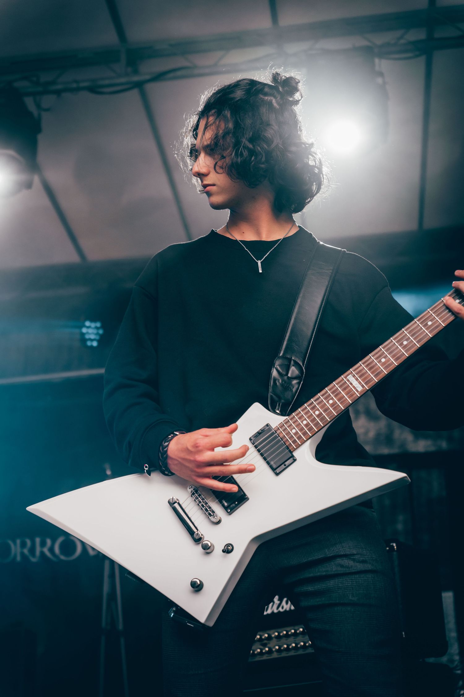 Long-haired guitarist plays white Explorer electric guitar on dimly lit stage with blue smoke.