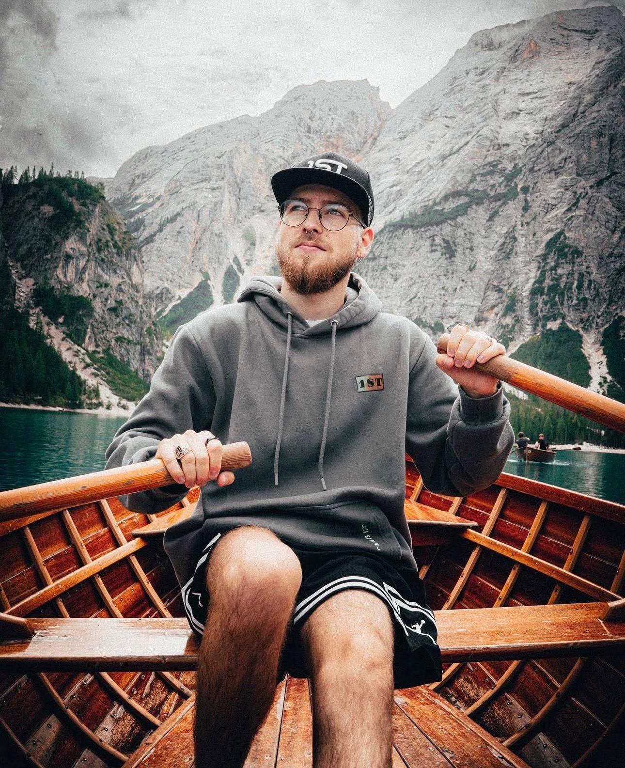 Bearded man in gray hoodie and cap rowing a wooden boat on a scenic mountain lake.