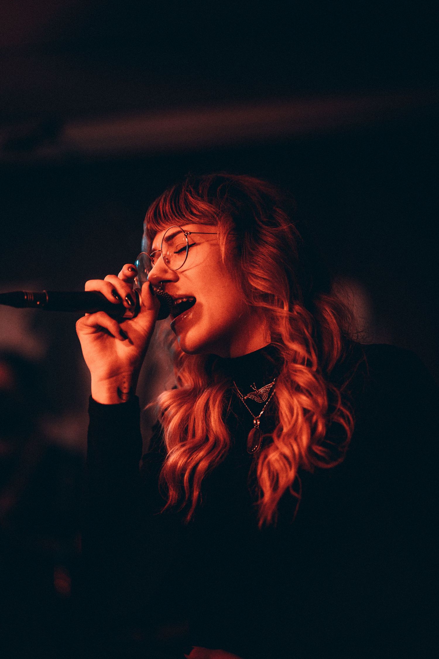 Female vocalist with glasses singing passionately under warm red stage lighting.