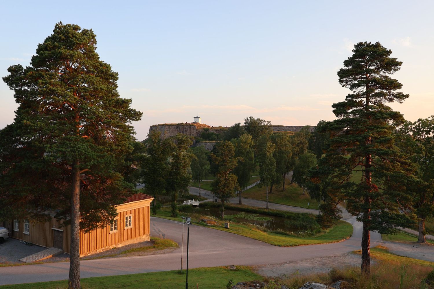 Scenic Scandinavian landscape with tall pine trees, red wooden building, winding path, and historic fortress on hill at golden hour.