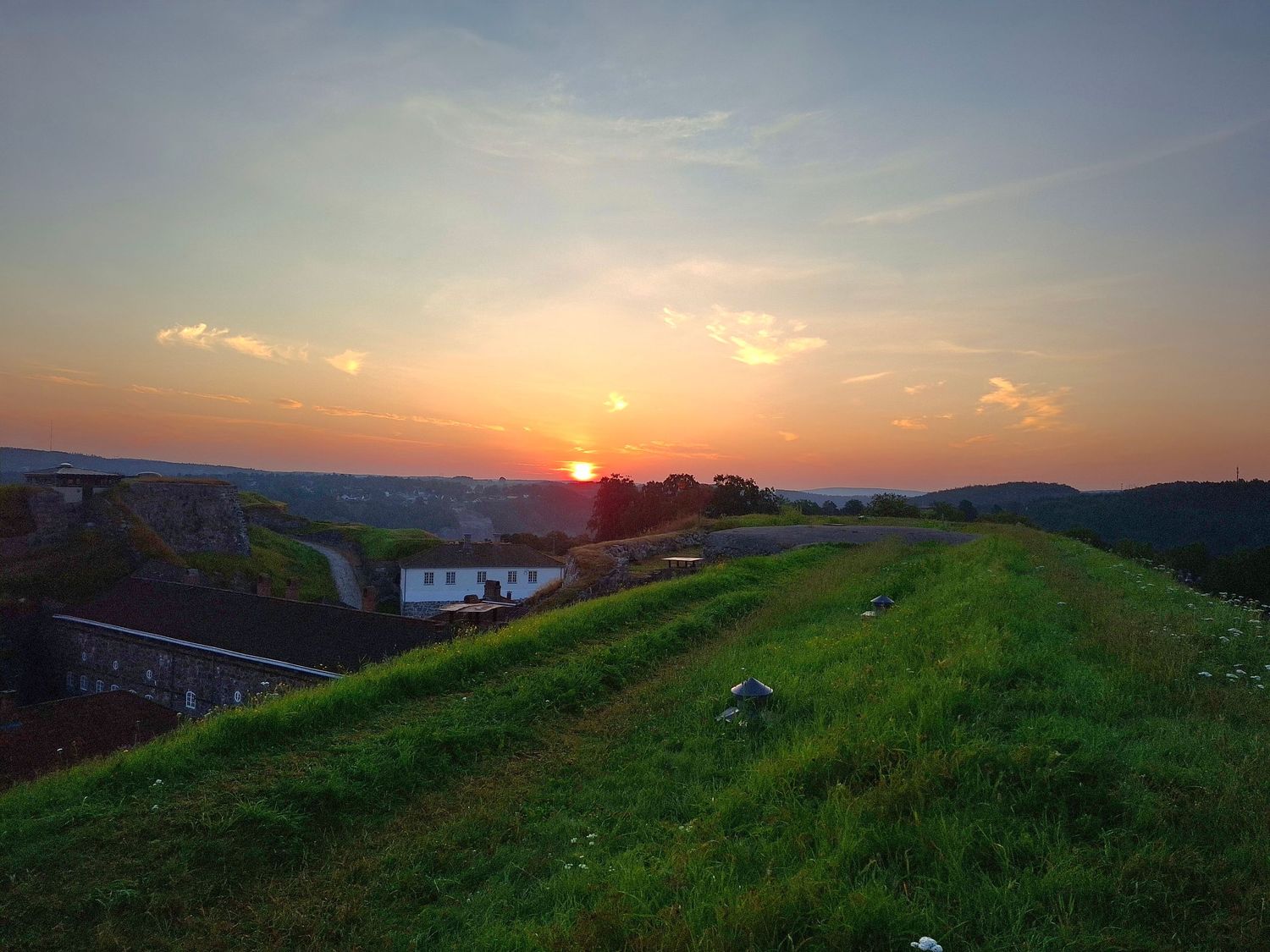 Stunning sunset over green rolling hills with a glowing orange sky and rural buildings in the background.