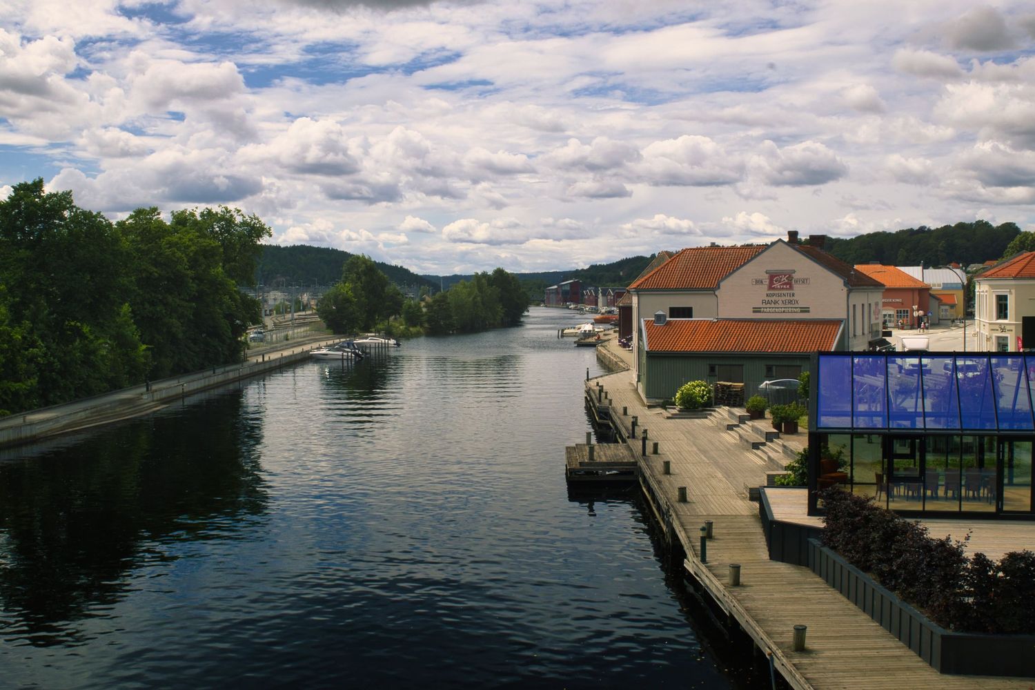 Scenic river canal with docked boats, waterfront buildings, and lush green trees under a partly cloudy sky.