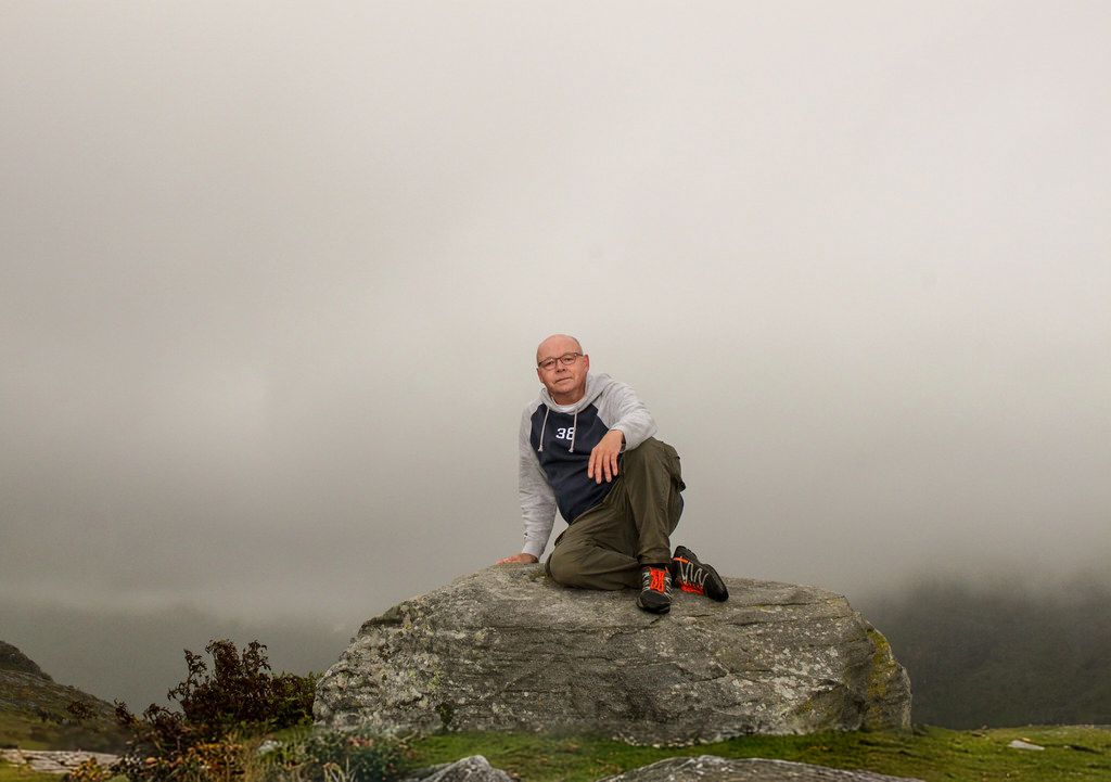 Bald man sitting on a rocky summit surrounded by thick fog and overcast skies in a mountainous landscape.
