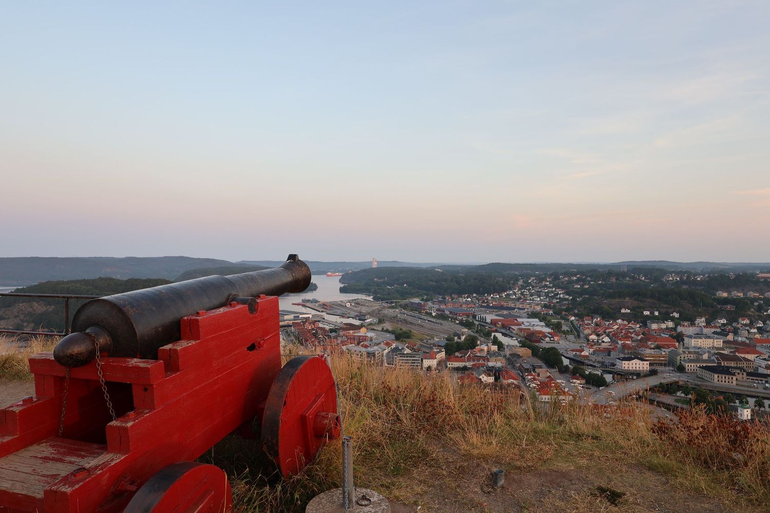 Red cannon on hilltop overlooks a coastal town and river at dusk, with a panoramic sky view.