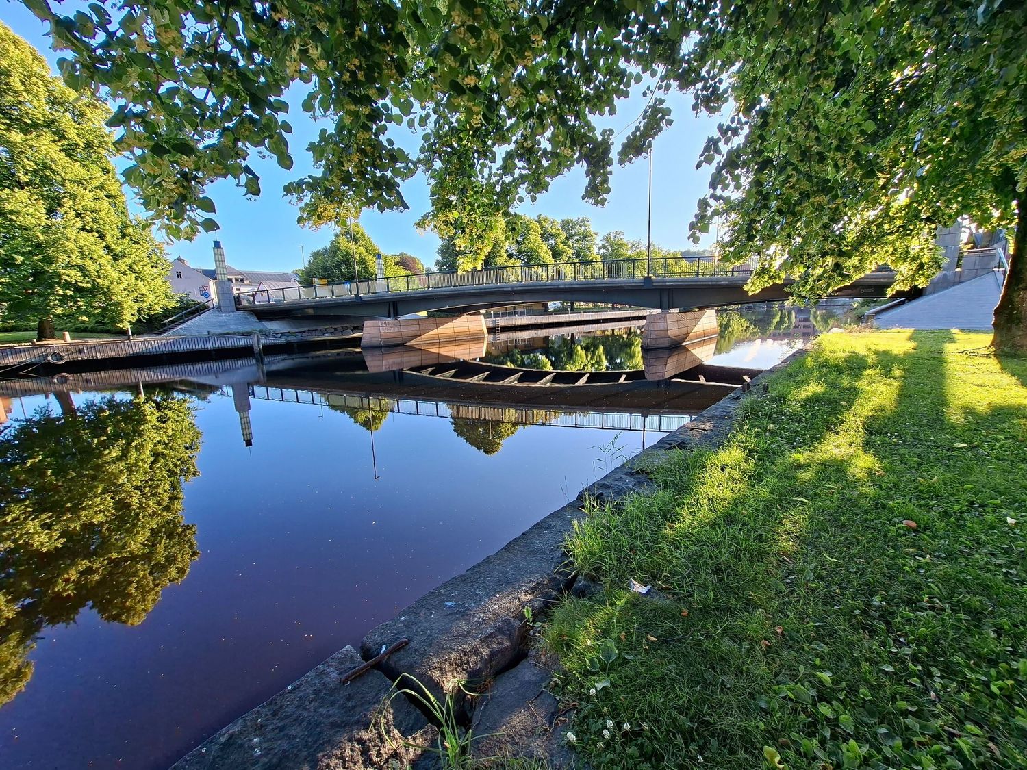 Serene canal with wooden boats moored under lush green trees beside a footbridge on a sunny summer day.