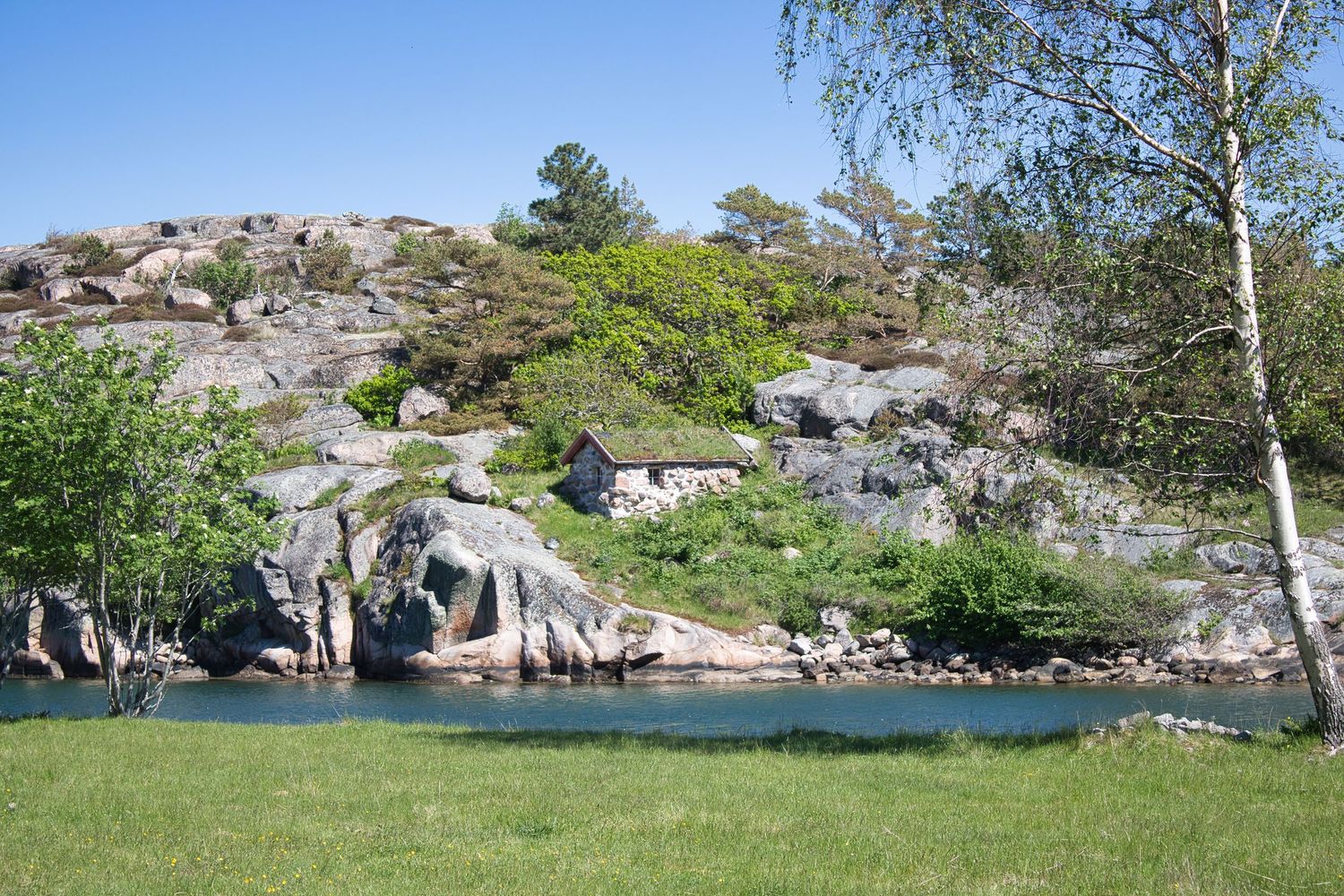 Rocky Scandinavian landscape with calm water, lush green trees, and granite boulders under a clear blue sky.