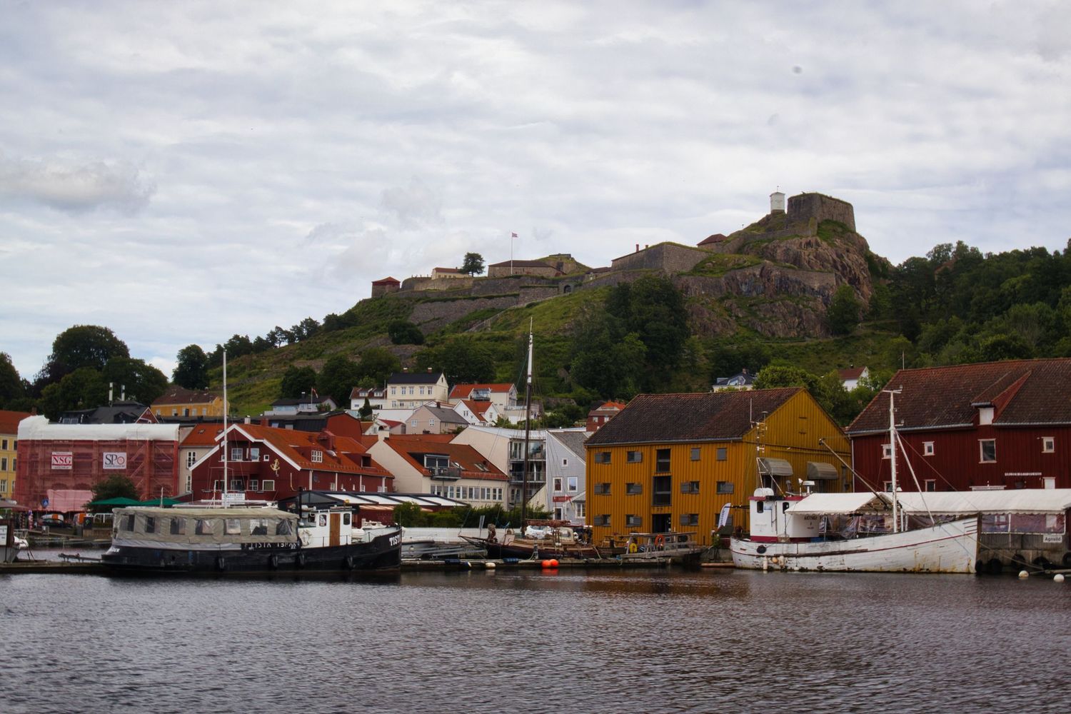 Colorful waterfront buildings and boats in a Norwegian harbor town with a historic fortress on a rocky hilltop.