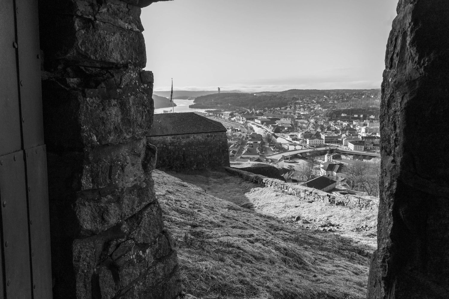 Black and white view of a coastal town through ancient stone castle walls, with a harbor visible in the distance.