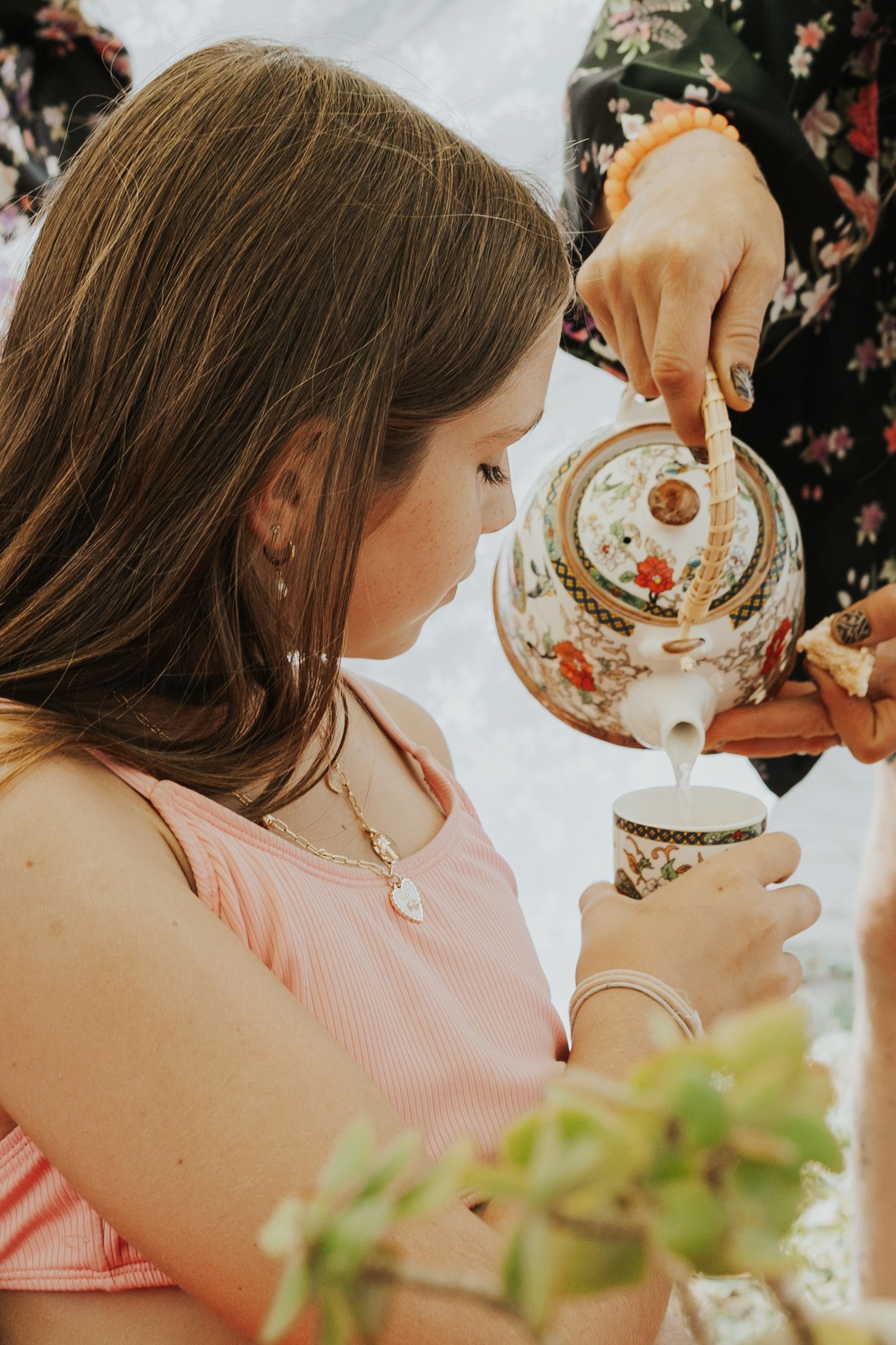 Girl in pink dress receiving tea poured from decorative floral teapot.