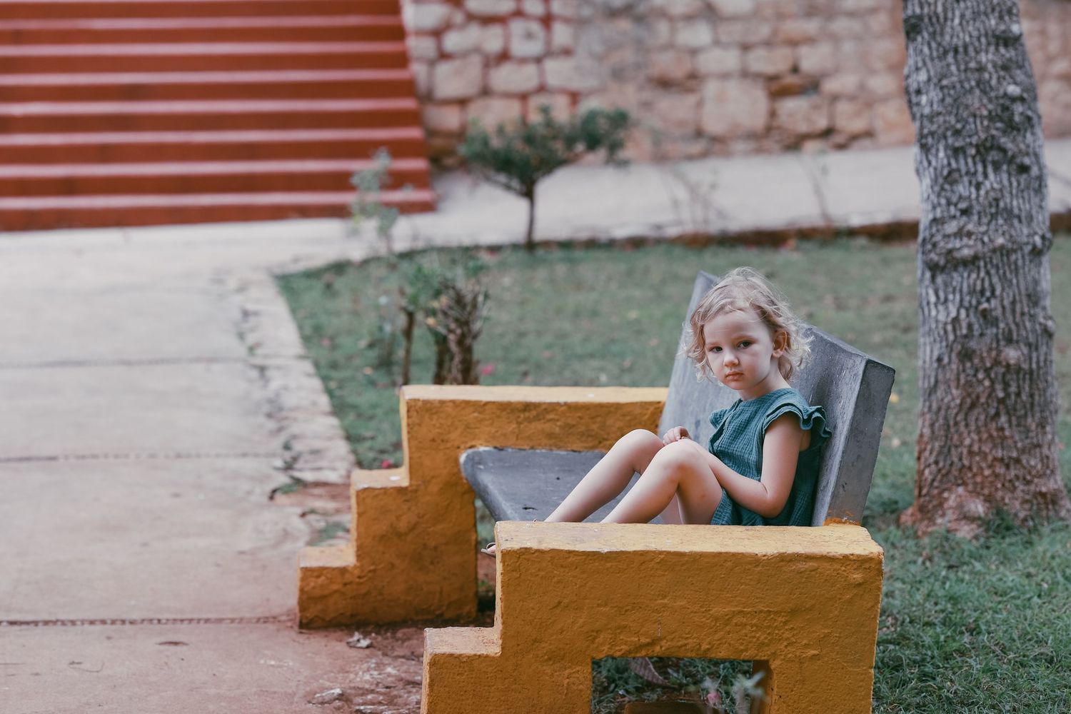 A young blonde girl in a teal dress sits alone on a yellow concrete bench outdoors.