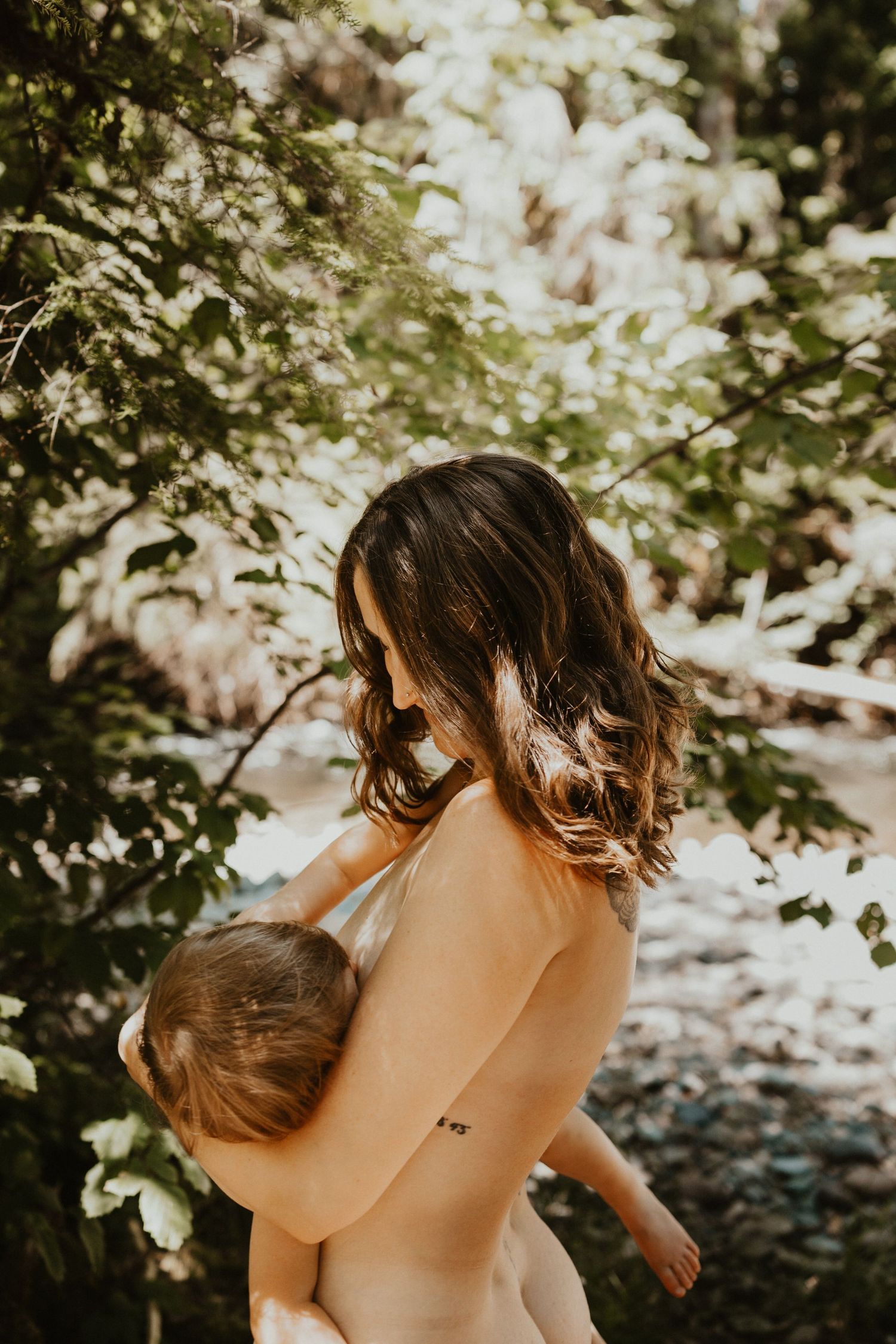Mother breastfeeding infant outdoors in dappled forest light.