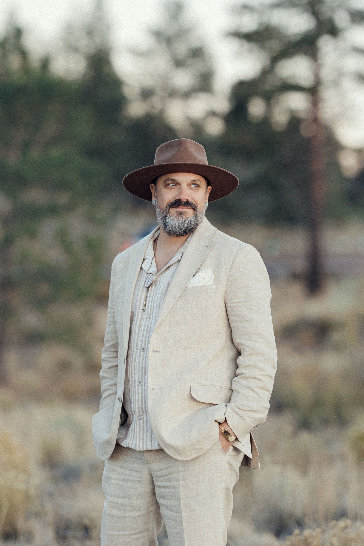 Bearded man in brown hat and linen suit standing confidently in outdoor forest setting.