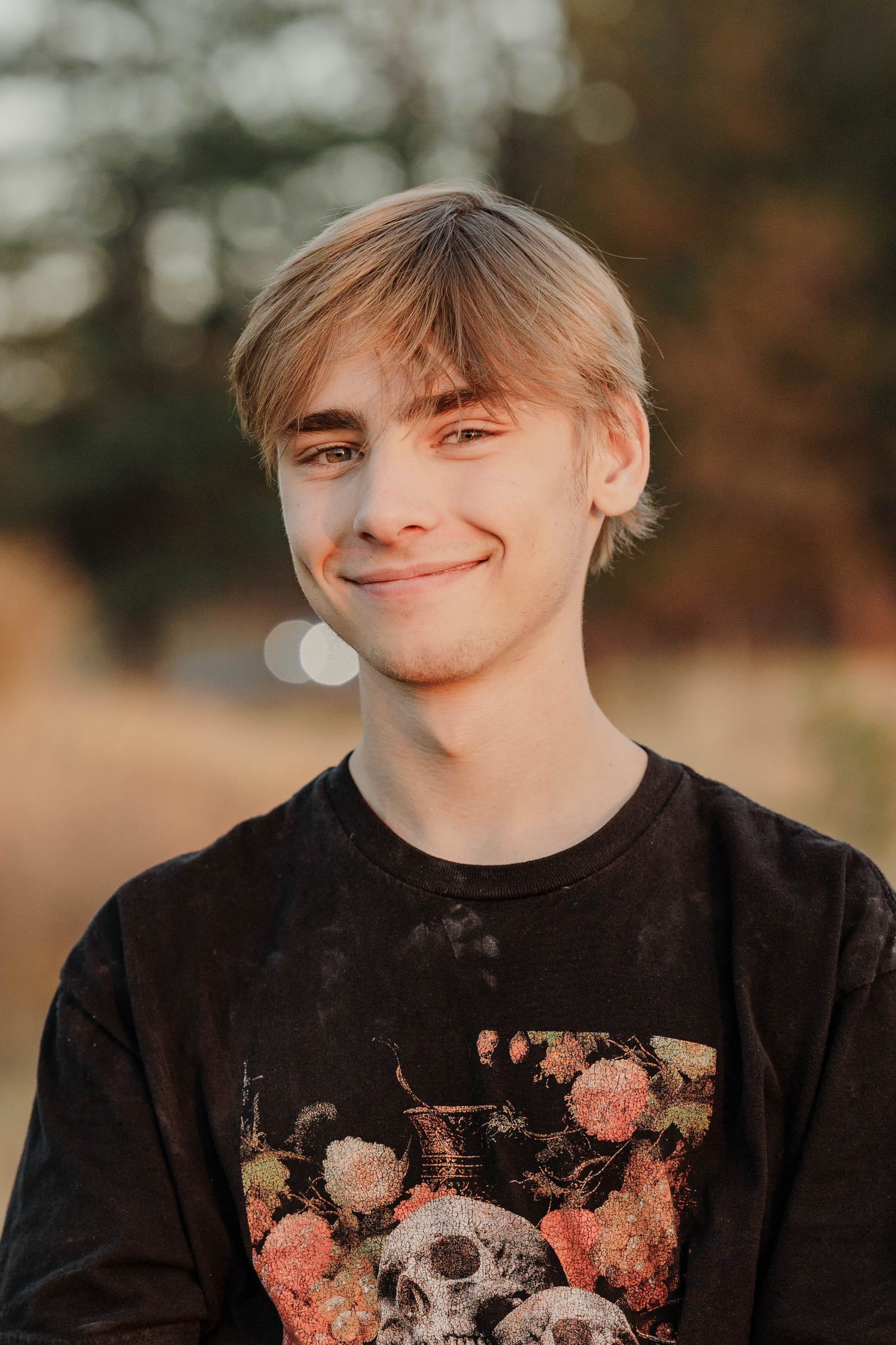 Teen boy with blonde hair smiling softly in skull floral graphic black tee outdoors.