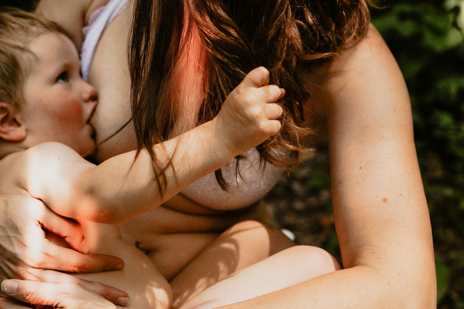 Mother breastfeeding her baby outdoors in natural sunlight, nurturing infant in a peaceful garden setting.