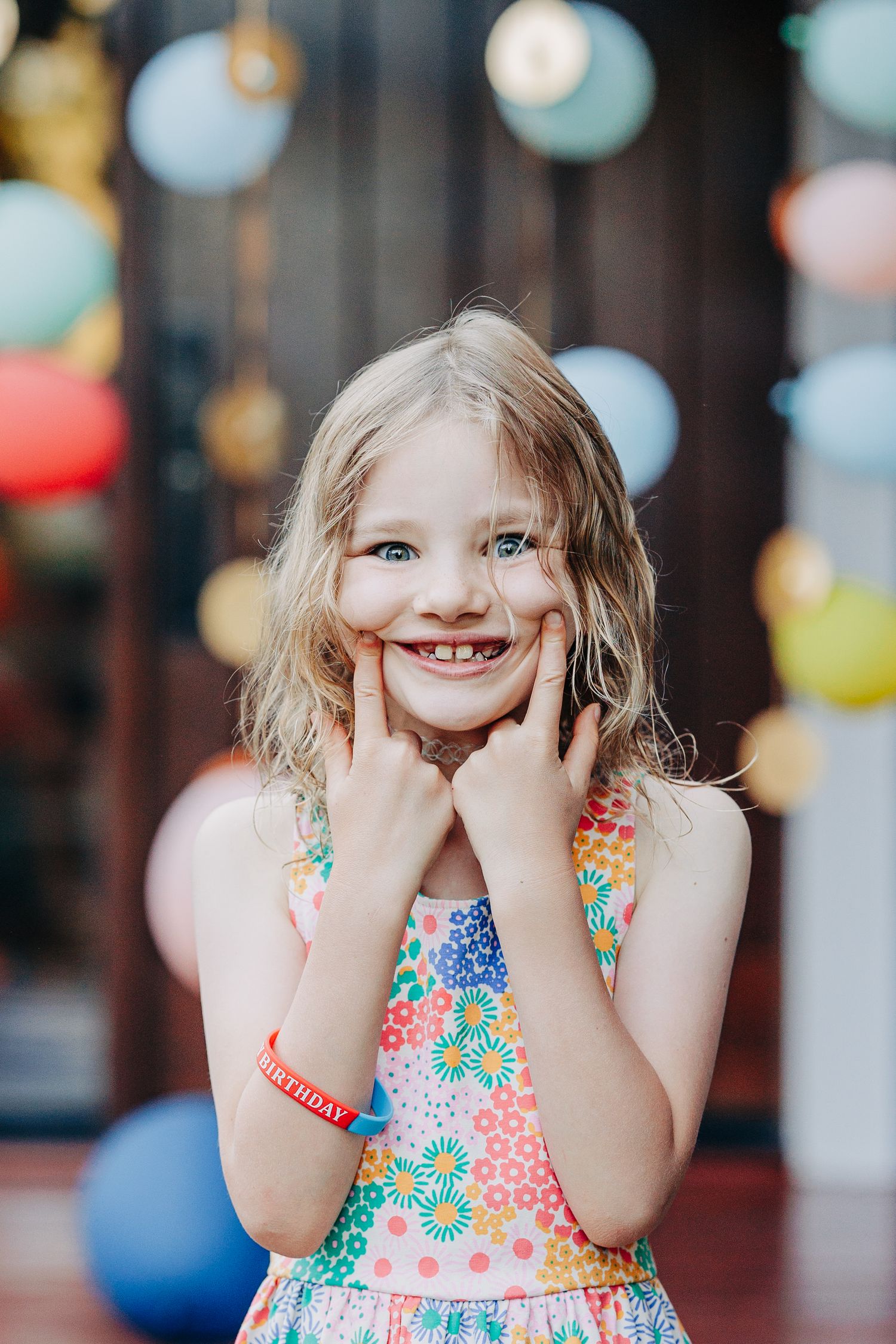 Happy birthday girl in floral dress makes big smile with fingers on cheeks surrounded by colorful balloons.
