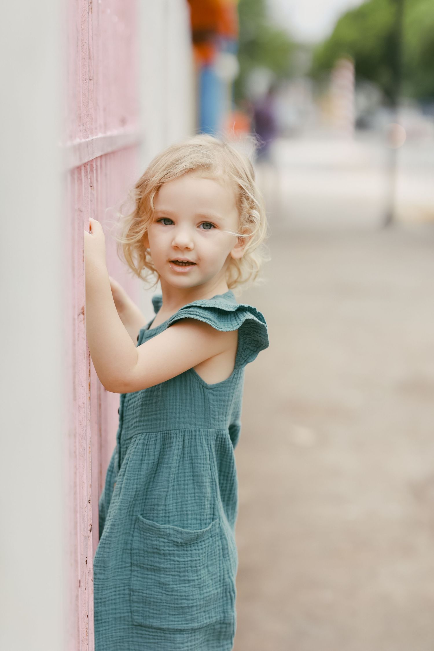 A blonde toddler girl in a teal dress leans against a pink fence post outdoors.