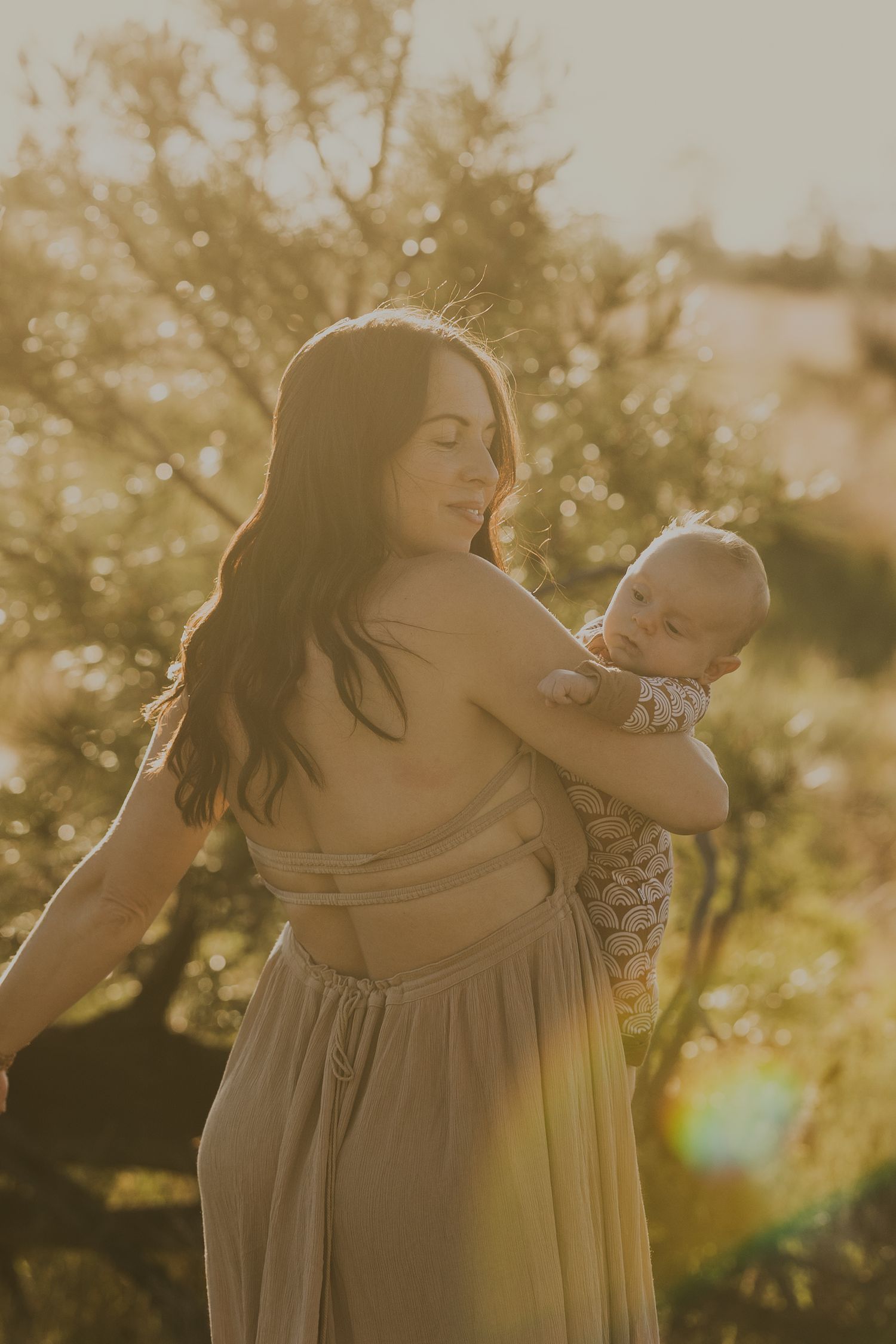 Mother in backless beige dress holding newborn baby bathed in warm golden sunset light outdoors.