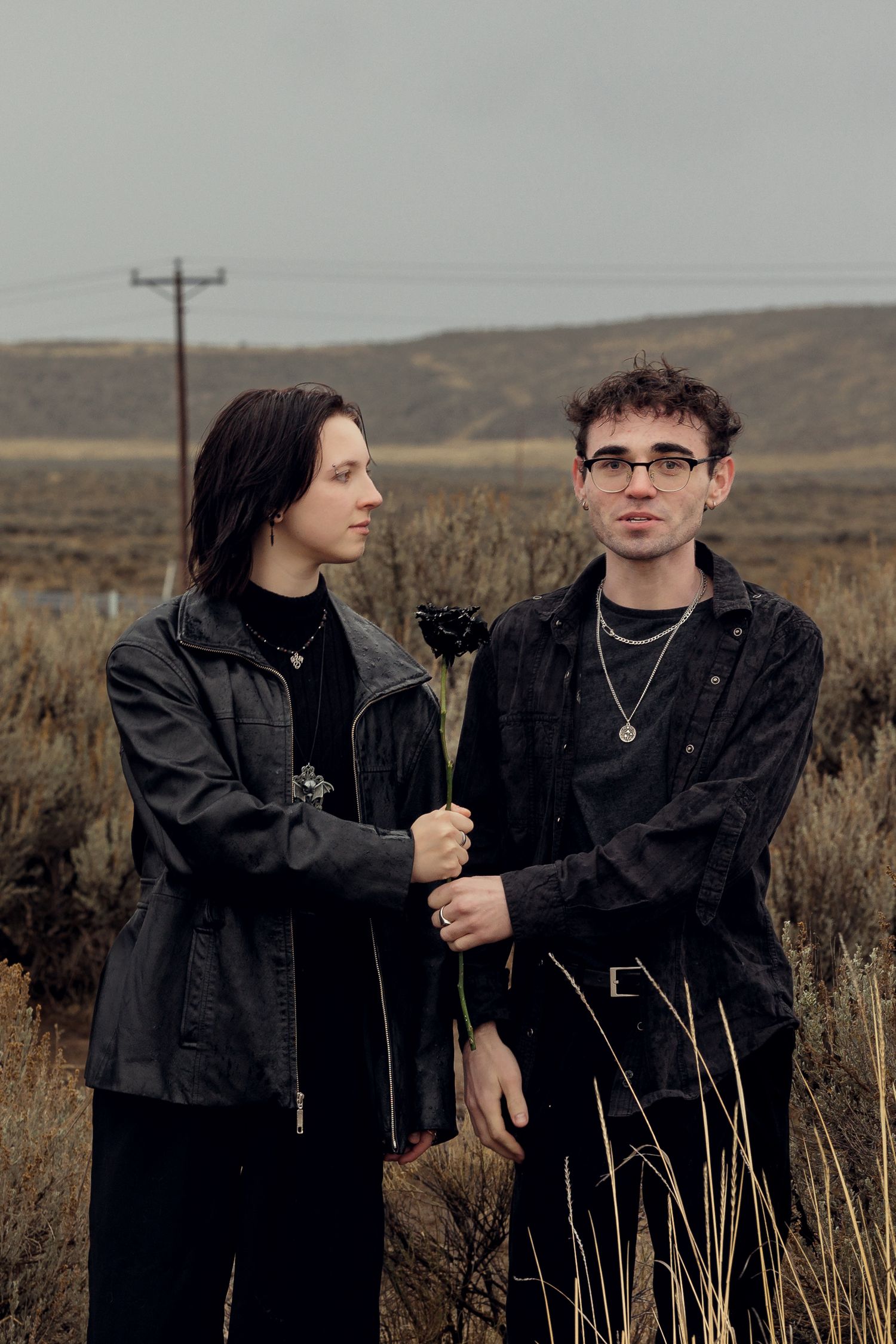 Two people in black clothing holding a dark flower together in a moody desert shrubland setting.