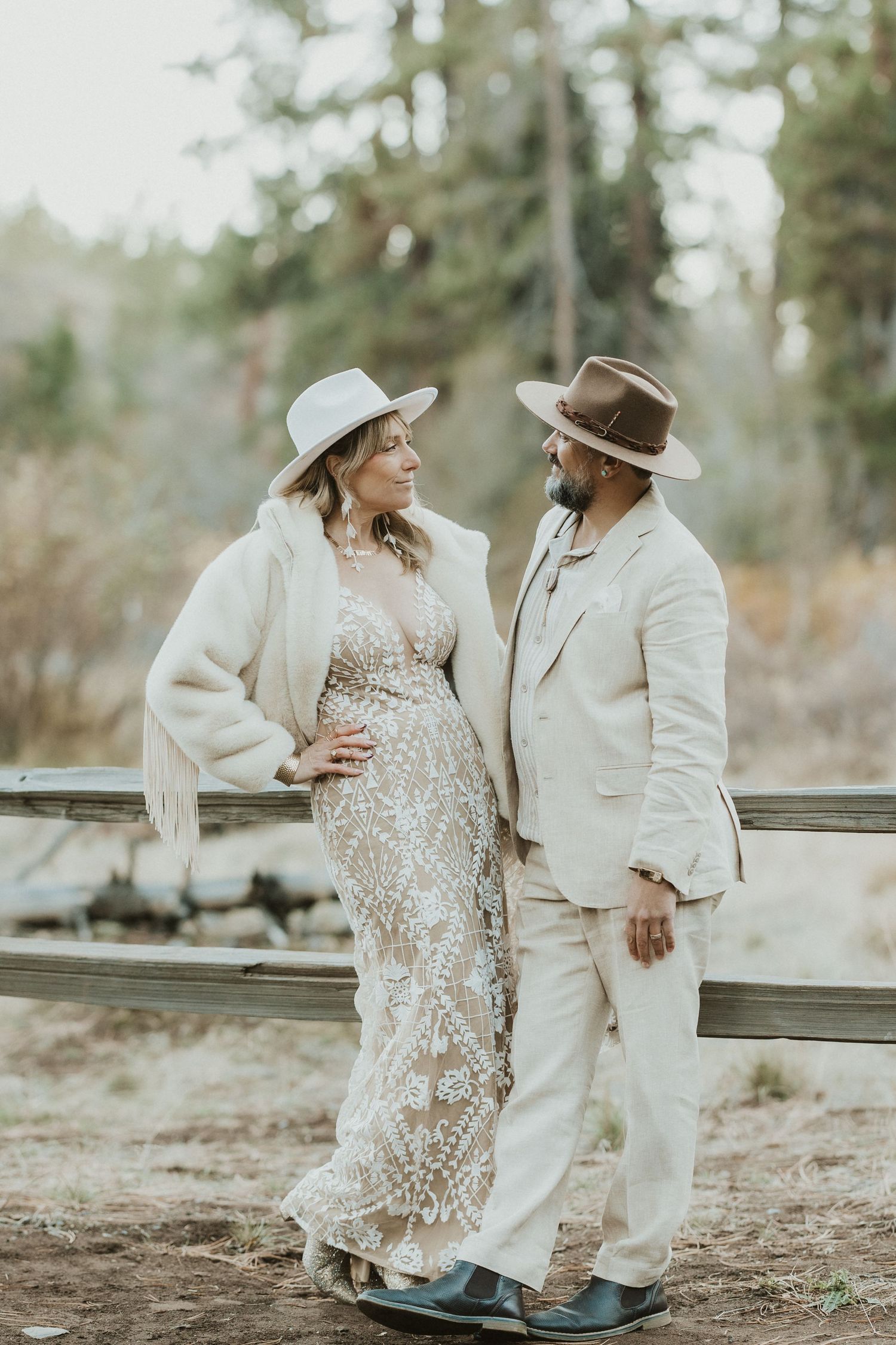 Pregnant couple in boho chic outfits and wide-brim hats posing by a rustic fence outdoors.