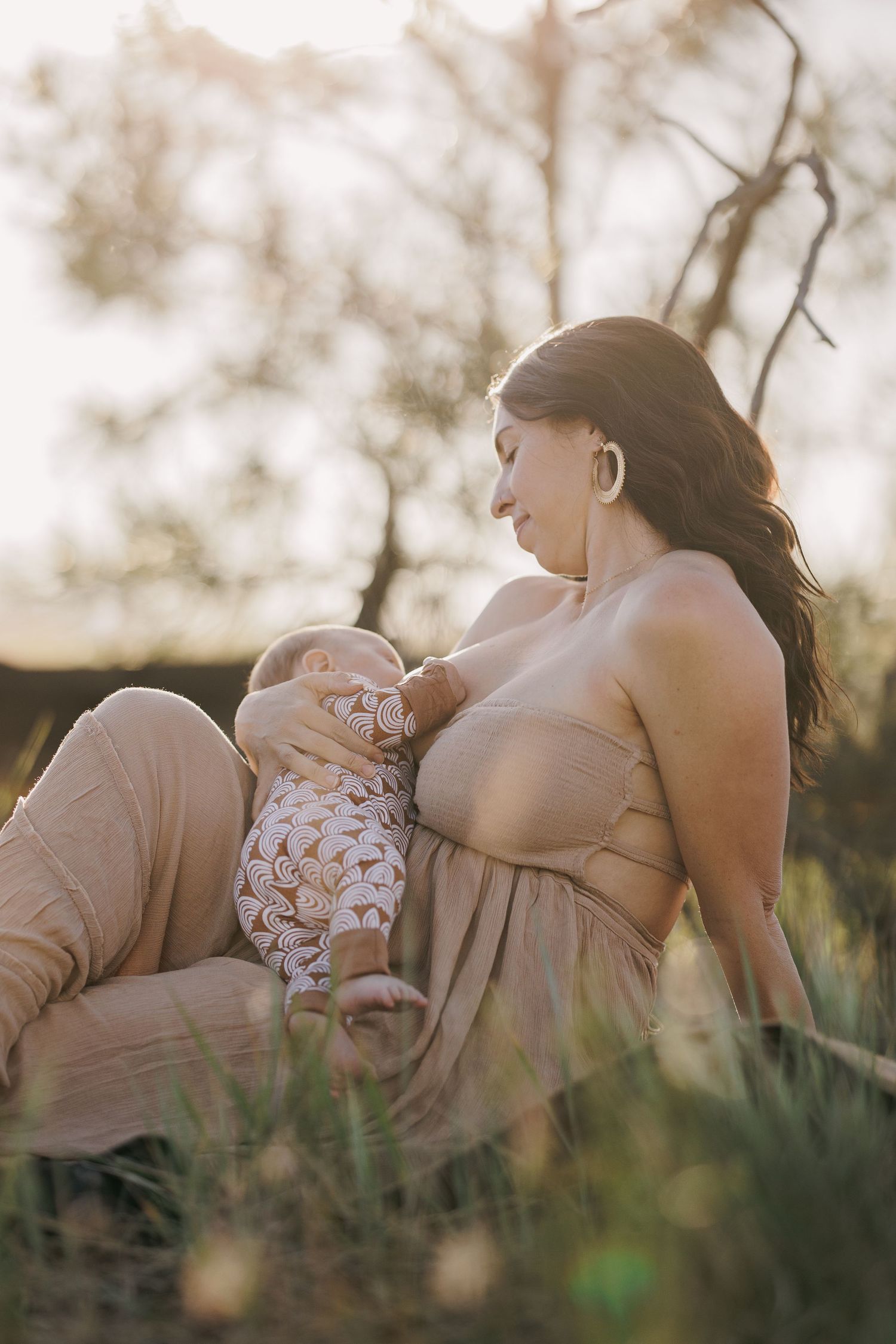 Mother in strapless beige dress breastfeeding newborn baby outdoors in warm golden sunset light.