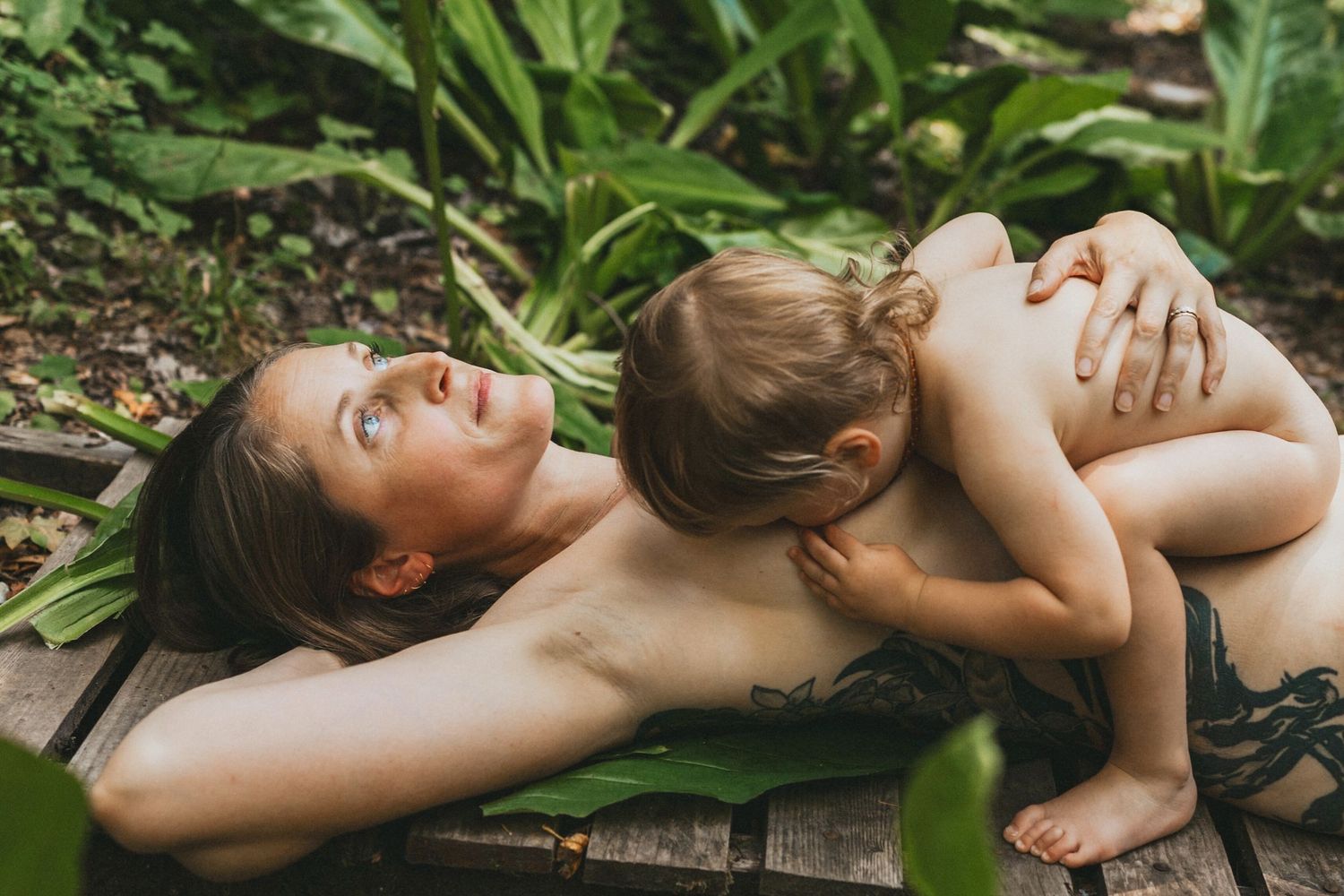 Mother and toddler bonding outdoors on wooden deck surrounded by lush tropical green plants.