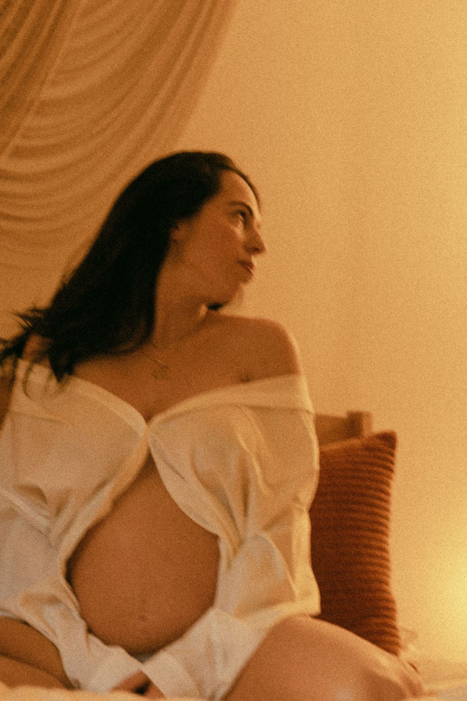 Pregnant woman in a white off-shoulder top sitting on a bed in warm golden light.