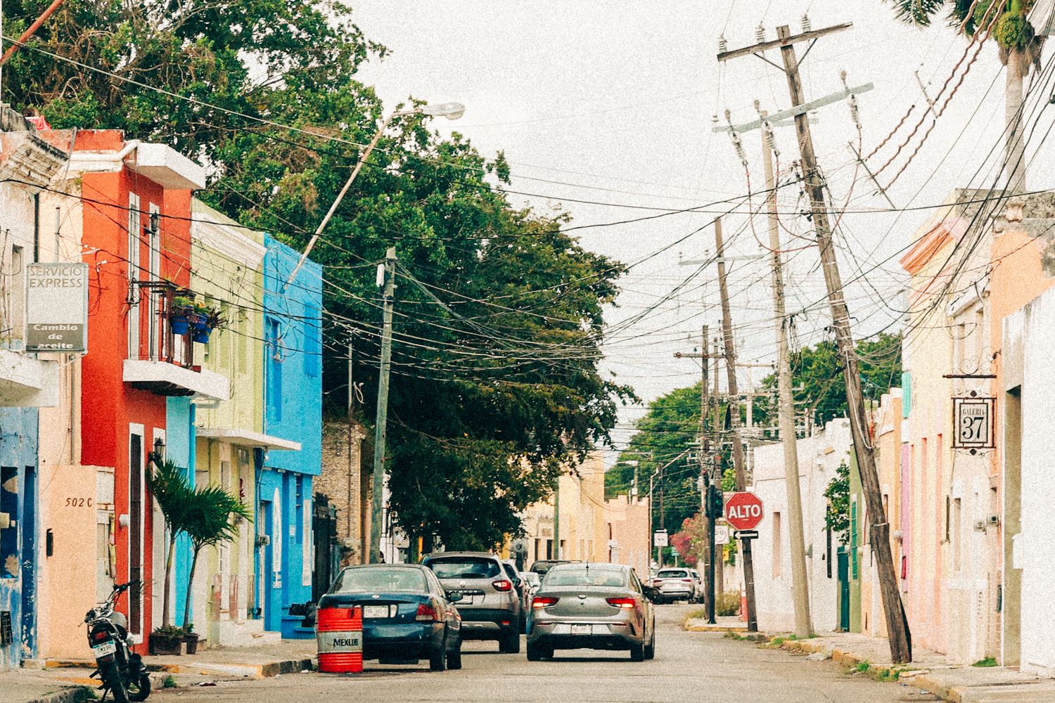 Colorful colonial street with cars parked along vibrant buildings and utility poles in a Latin American town.