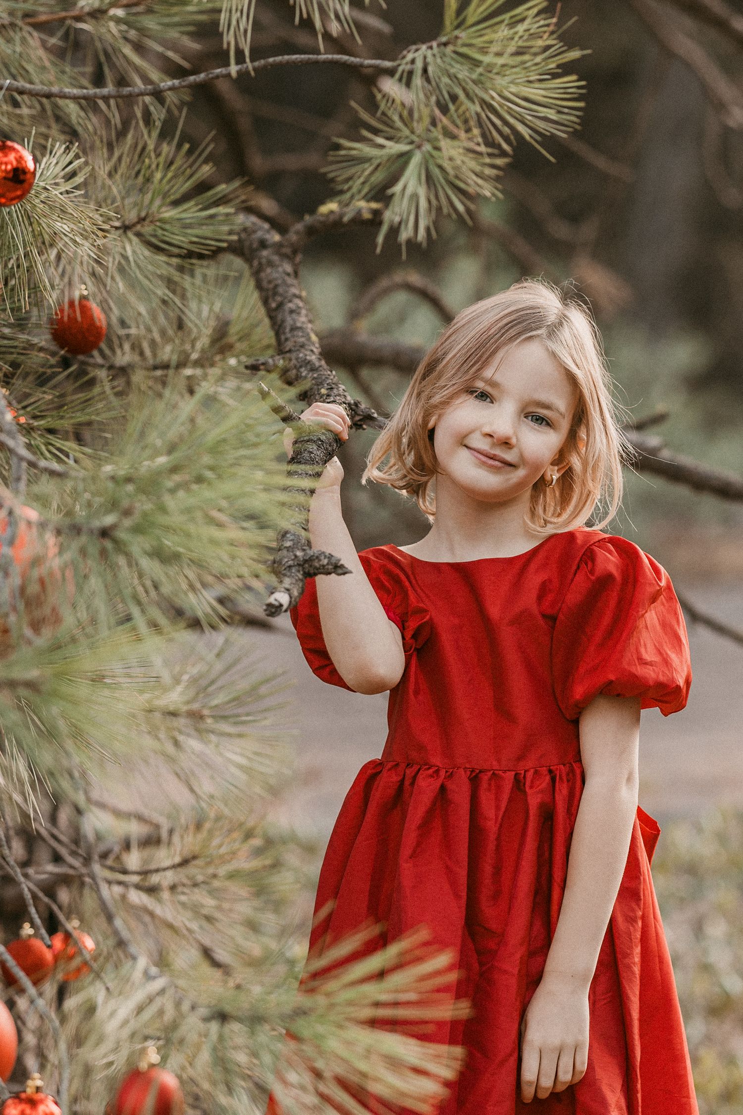 Blonde girl in red dress standing beside pine tree decorated with red Christmas ornaments in forest.